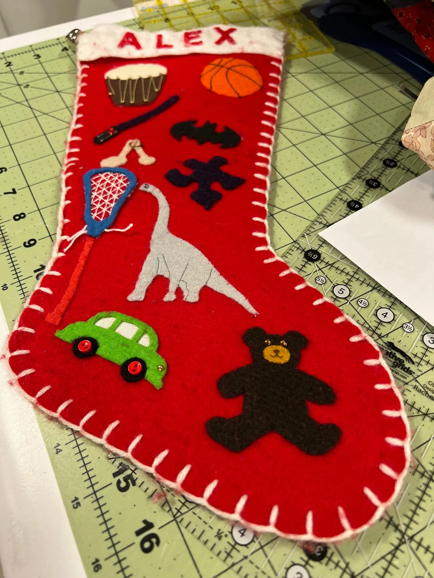 A red felt Christmas stocking edged in white blanket stitch, with ALEX appliquéd across a white cuff, and shapes down the front including a drum, a basketball, a lacrosse stick, a hot-air balloon, a gray dinosaur, a green car and a brown teddy bear.