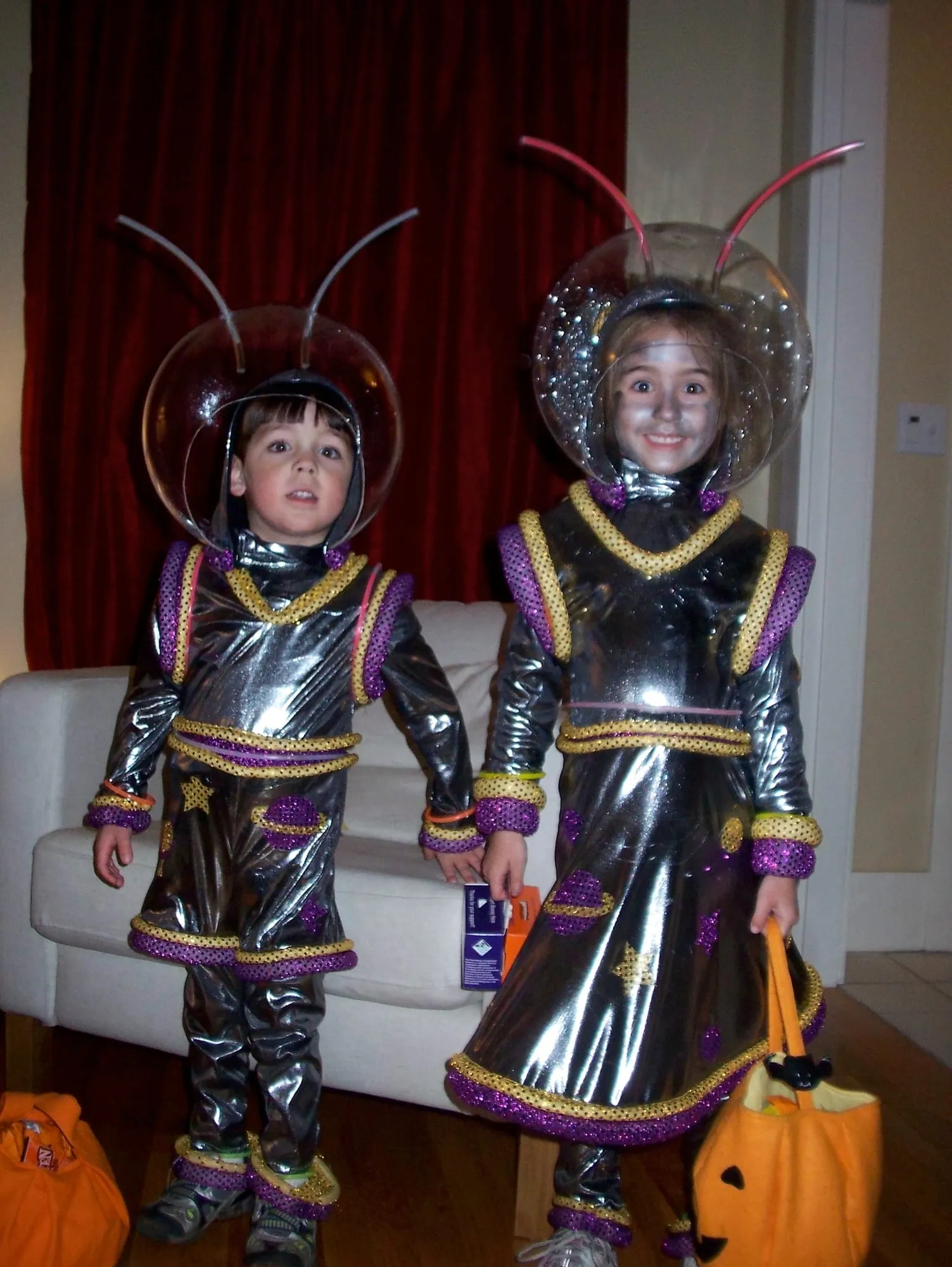 Two children in silver alien costumes with clear domed helmets and pink antennae, trimmed in gold and purple sequins, holding trick-or-treat bags.
