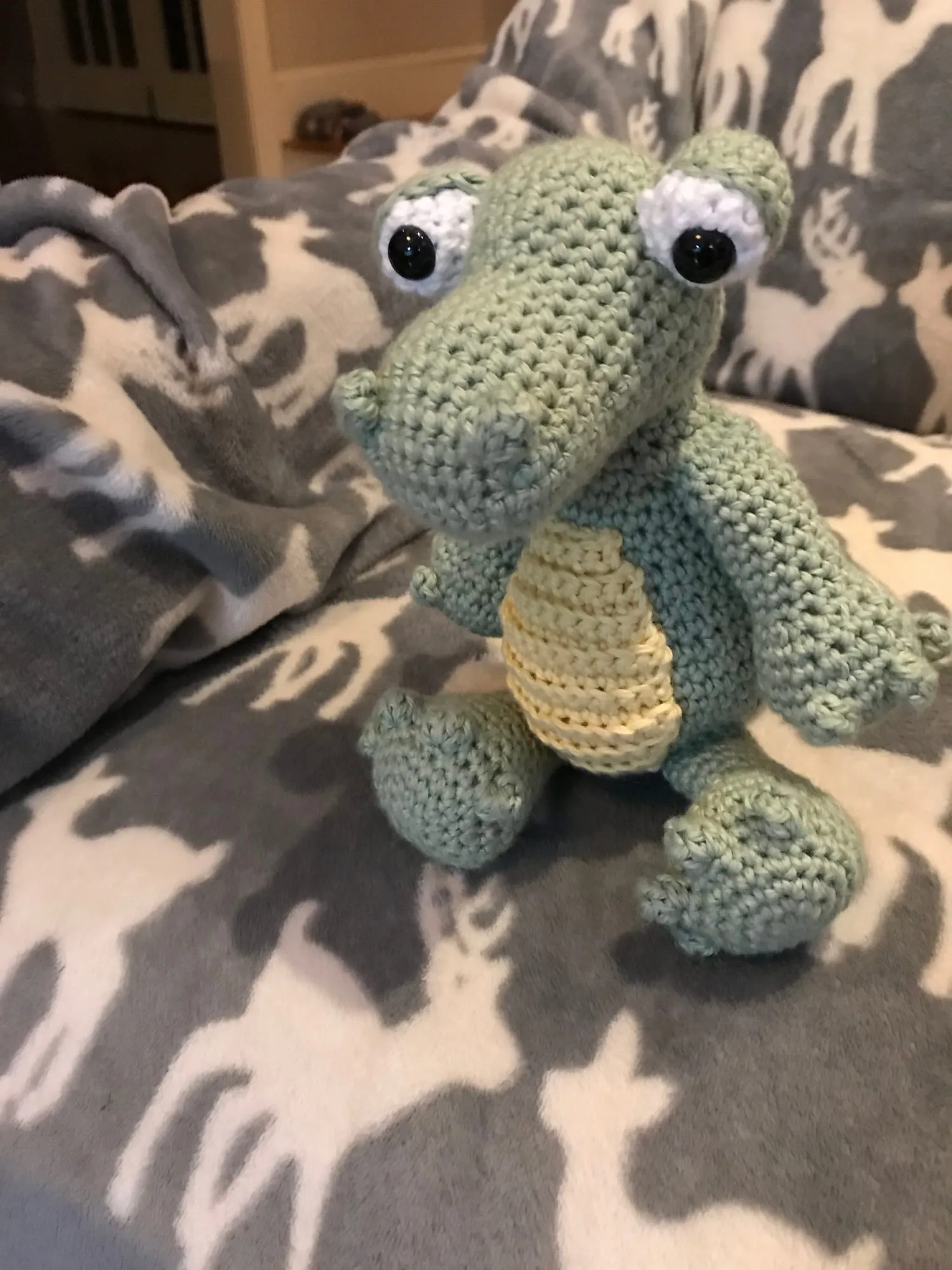 A crocheted alligator sitting upright in two greens, with a pale belly, round white eyes and a long snout.
