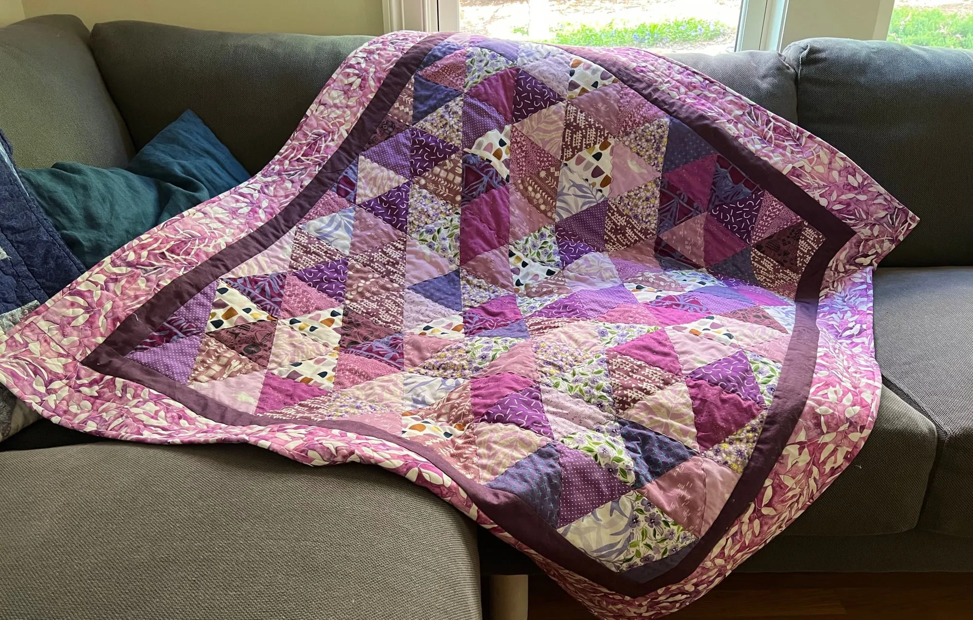 A baby quilt draped over the arm of a sofa, pieced in triangles of purple, lilac and pink prints inside a floral border.