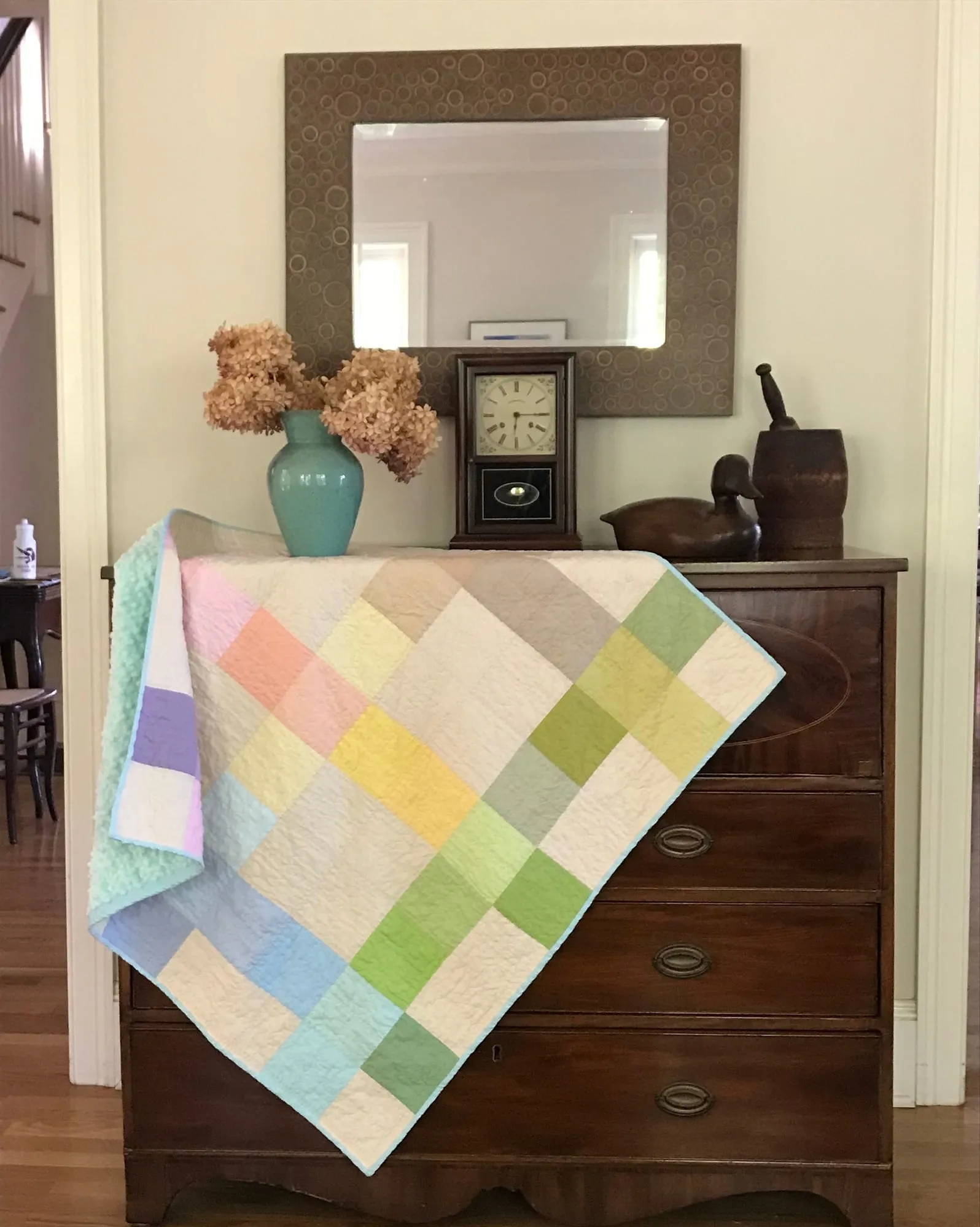 A baby quilt in pastel squares of green, blue, yellow and pink, folded over the corner of an antique chest of drawers.