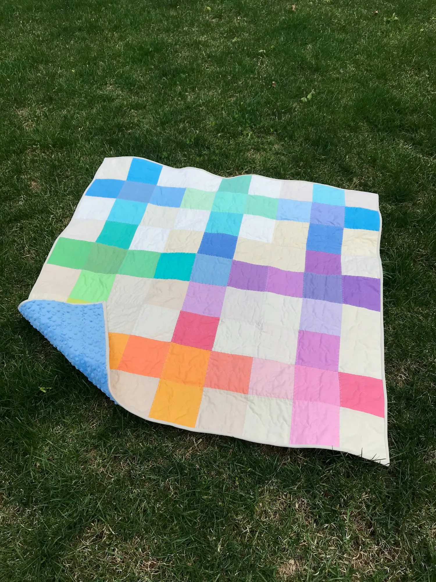 A baby quilt spread on grass with one corner turned back to show a blue dotted backing, its front a grid of large pastel squares in blue, green, pink, purple and orange inside a cream border.
