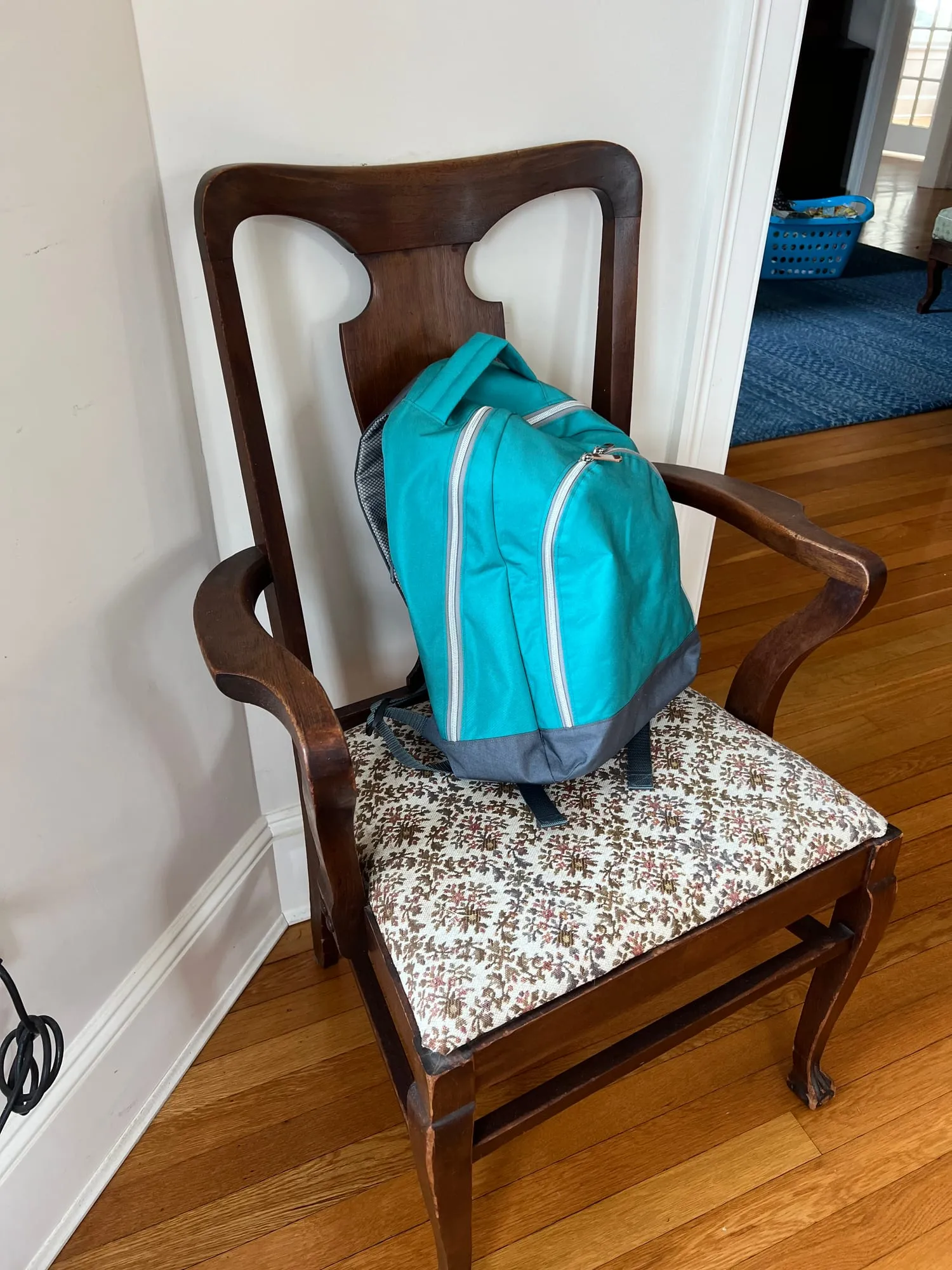 A small turquoise backpack with a curved zip and a front pocket, standing on the seat of a wooden chair.