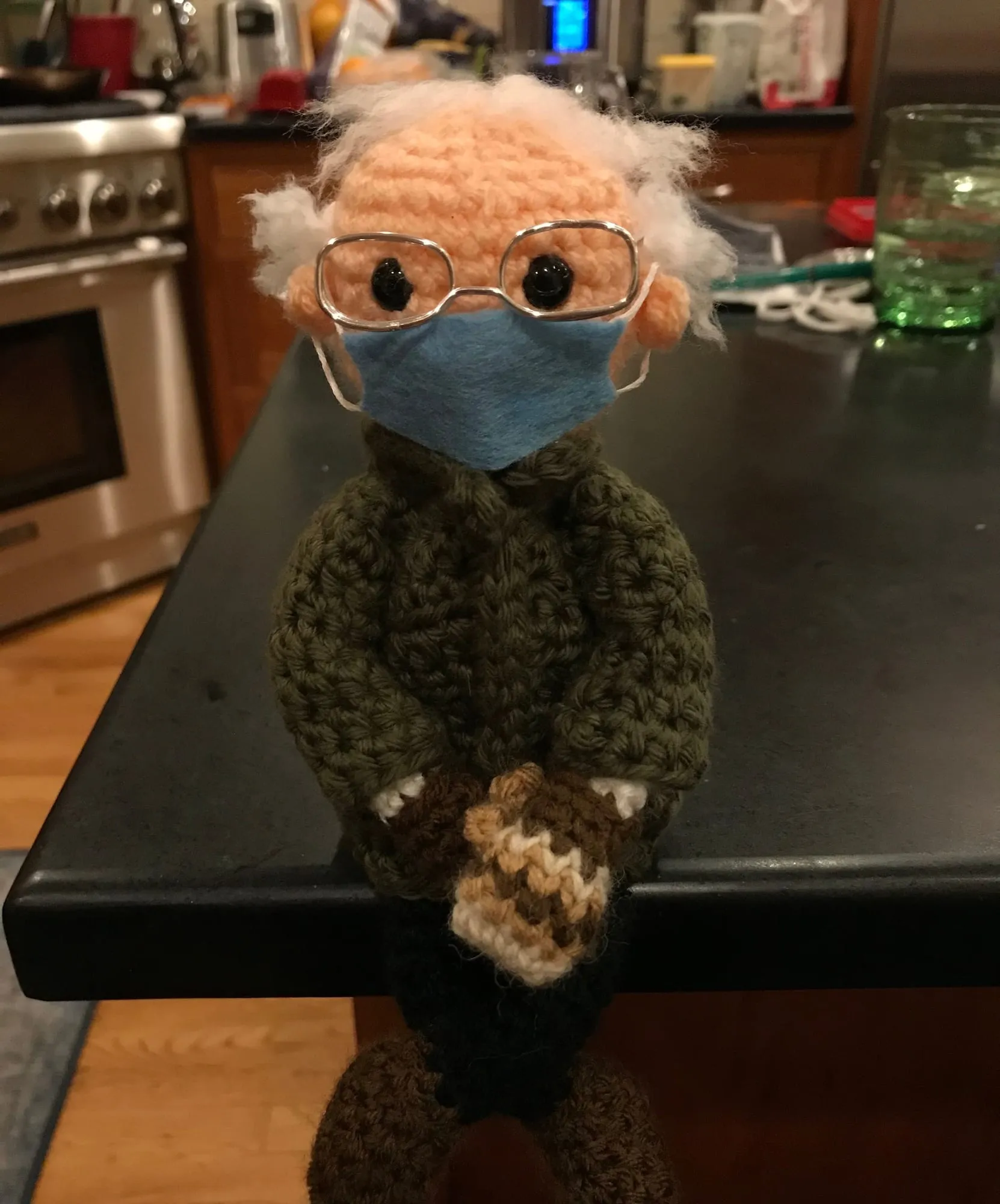A crocheted figure of Bernie Sanders propped at the edge of a kitchen counter, in an olive coat, a blue face mask, round glasses and patterned mittens, with a shock of white hair.