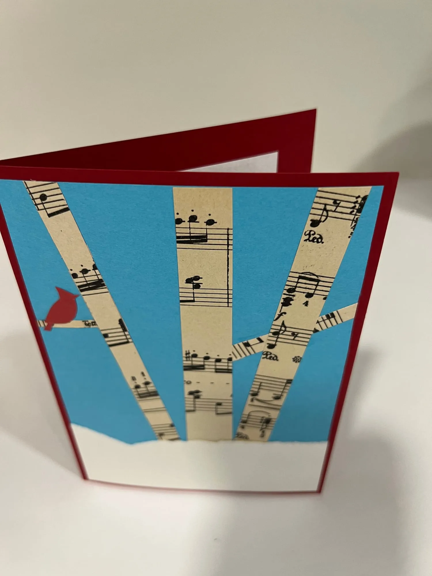 A folded card standing open, its front showing three birch trunks cut from printed sheet music against a bright blue sky, with a small red cardinal perched on one.