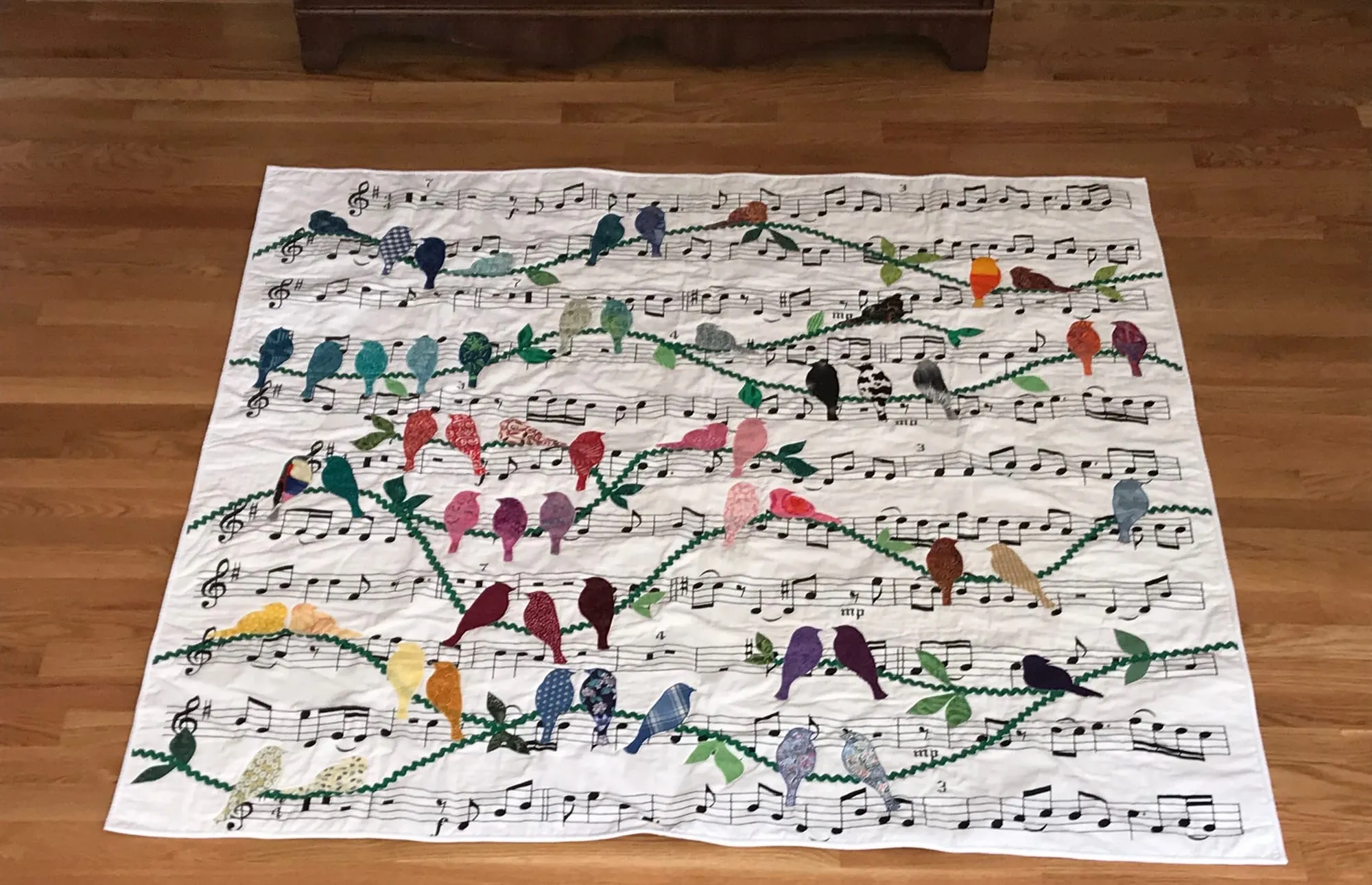 A quilt laid on a wooden floor, printed with staves of sheet music and appliquéd with rows of small birds in many colors perched along the lines.