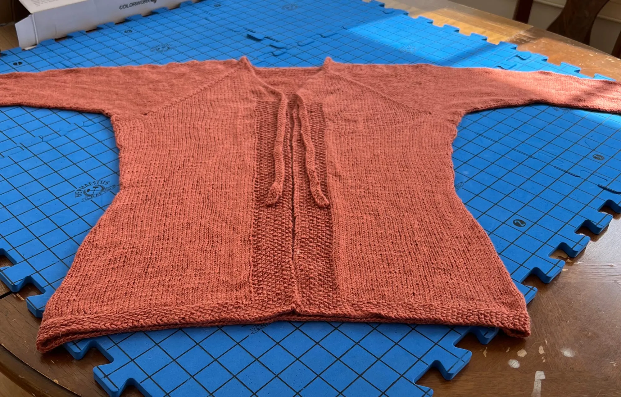 A coral cardigan laid open on blue blocking mats to dry, with long sleeves and ties at the waist.