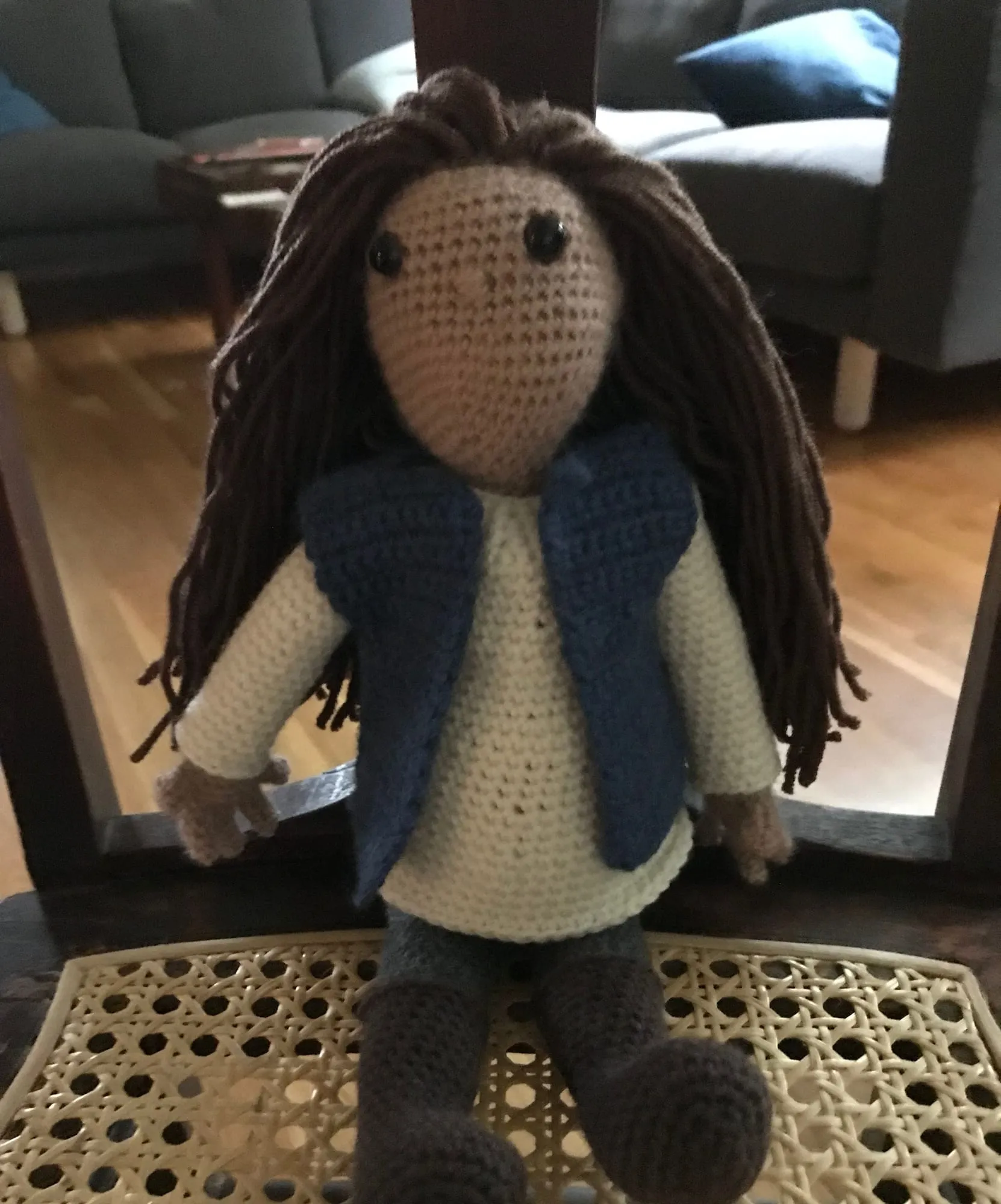 A crocheted doll with long brown hair, wearing a cream top, a blue vest and dark trousers and shoes.