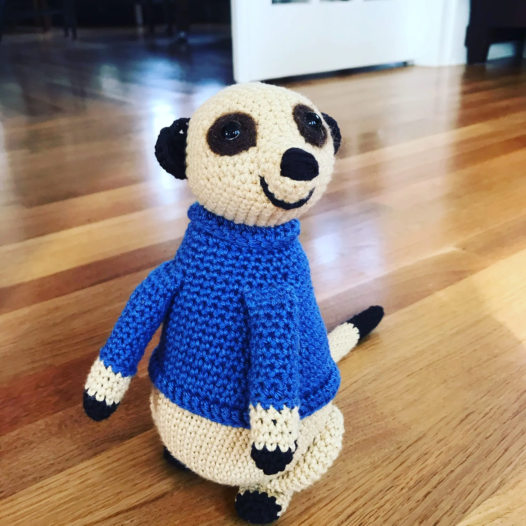 A crocheted meerkat standing upright in a blue sweater, with a cream body, dark eye patches, dark paws and a long tail behind.
