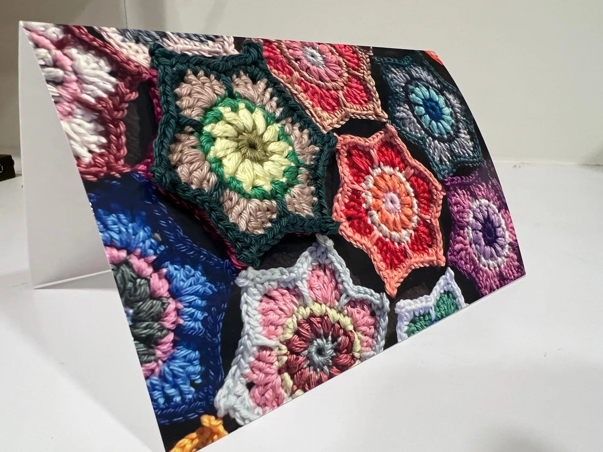 A folded card standing on a white surface, its front a photograph of crocheted hexagon ornaments printed at their real size, with one actual crocheted ornament fixed over its own picture.