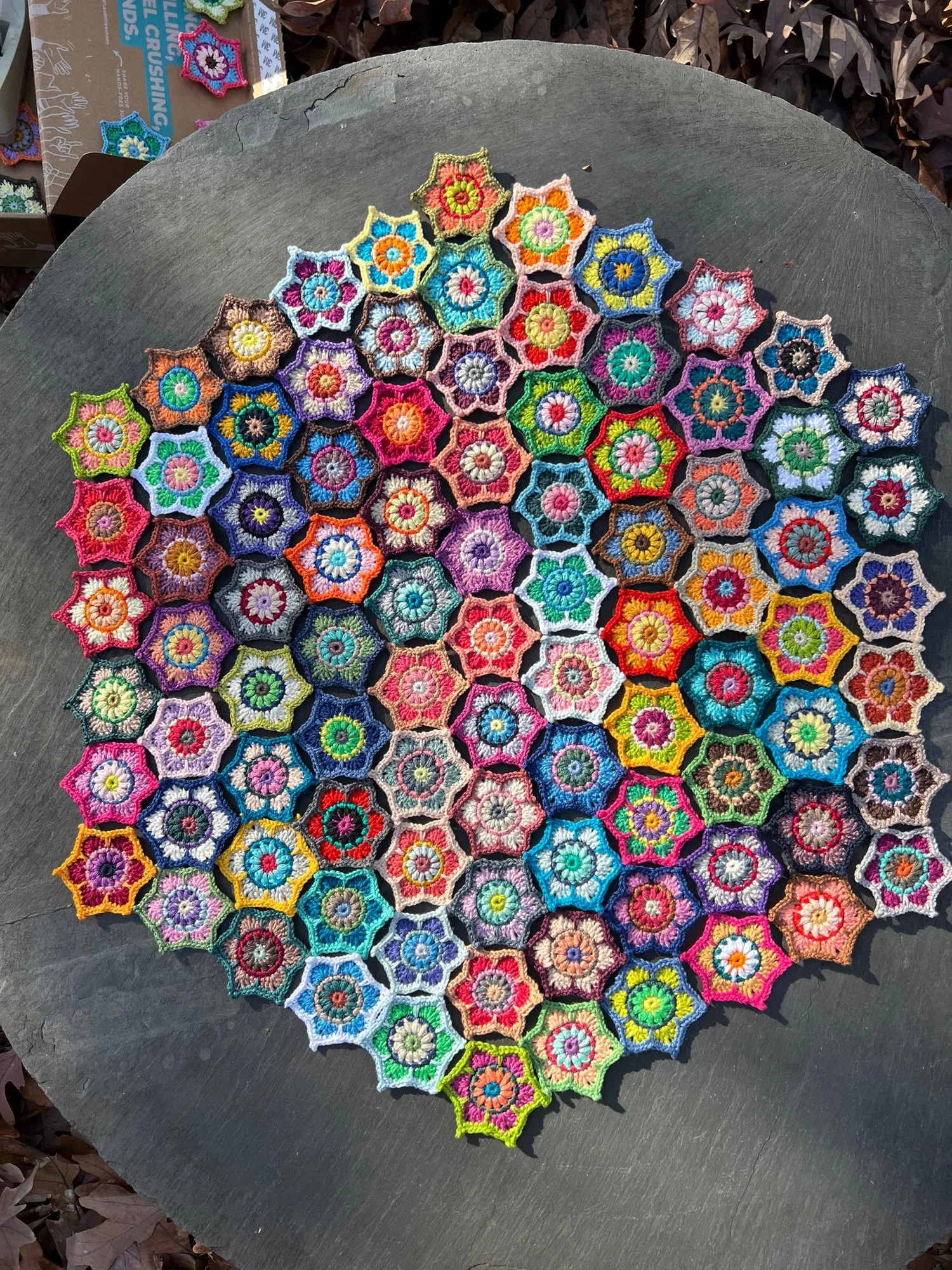Dozens of crocheted hexagon ornaments laid out on a round table outdoors, arranged edge to edge into one large hexagon, each worked as a flower in a different combination of colors.