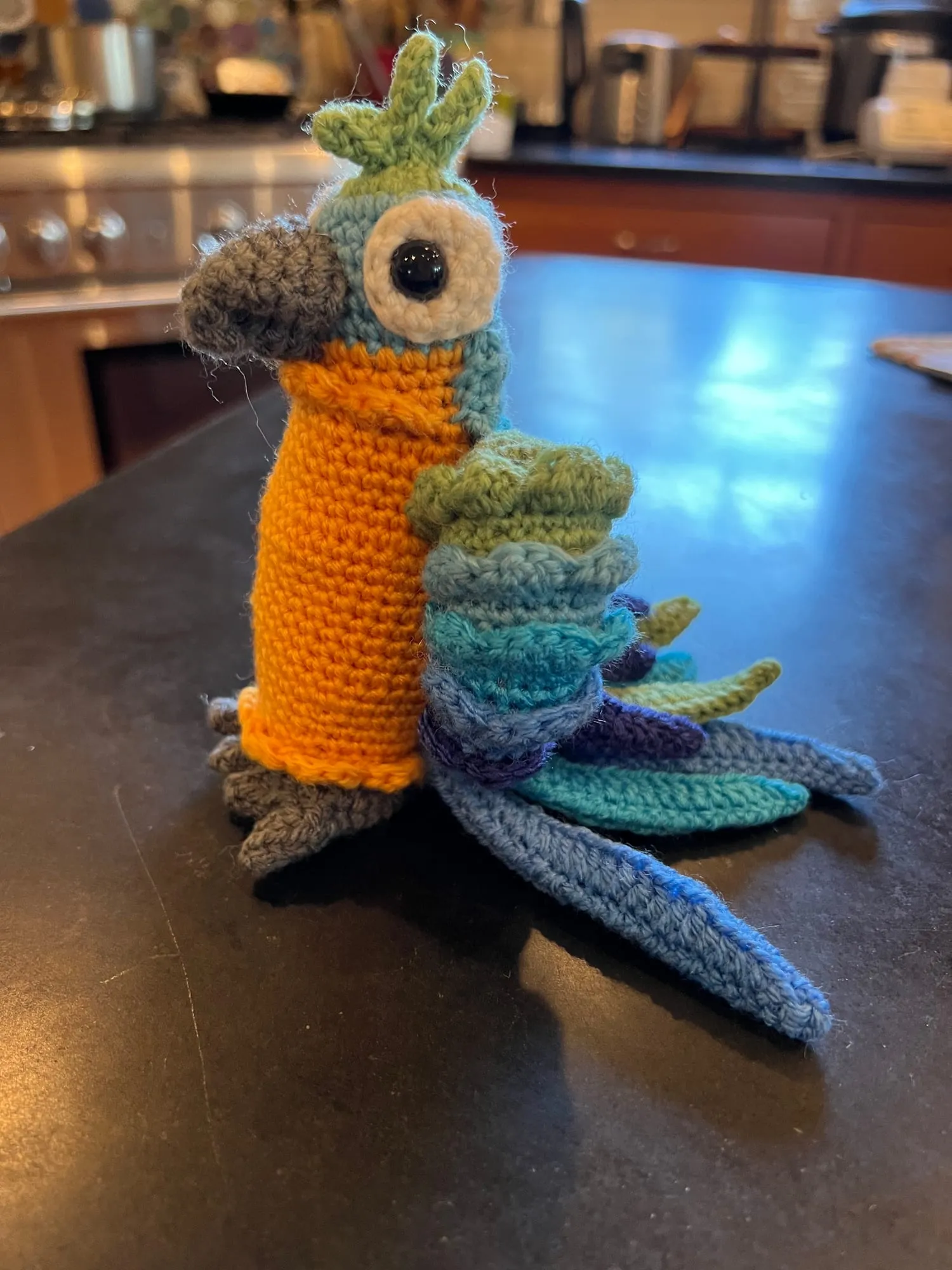 A crocheted parrot with a bright orange body and a long layered tail in green, teal and blue, standing on a dark tabletop.