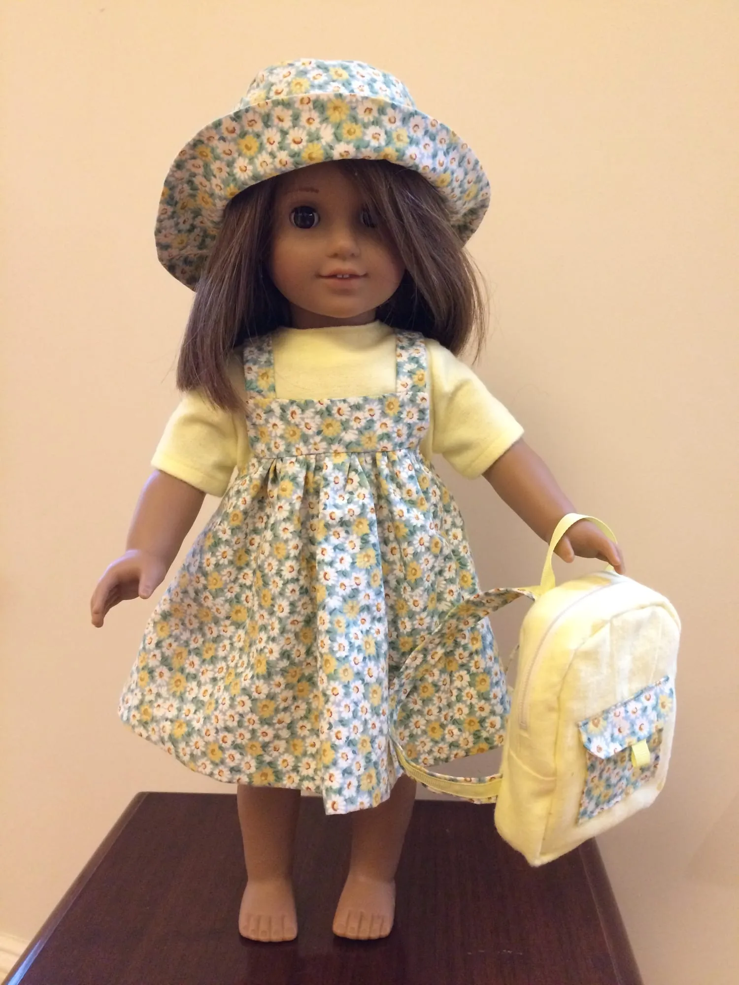 An eighteen-inch doll in a floral pinafore dress over a pale yellow top, with a matching brimmed hat and a small backpack.