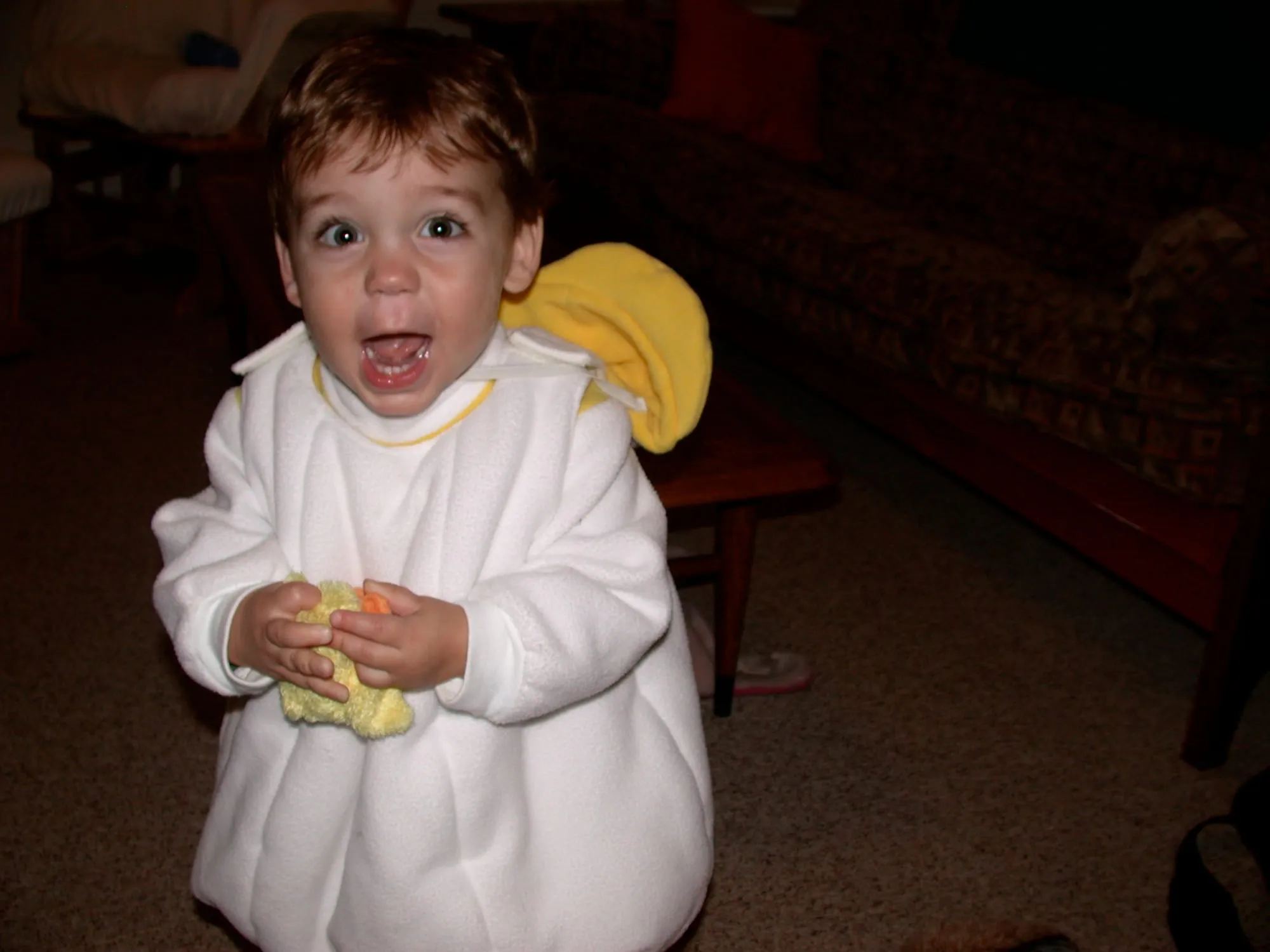 A toddler in a white duck costume, a wide smock with a yellow-trimmed collar and a yellow bill hood pushed back behind the shoulder.