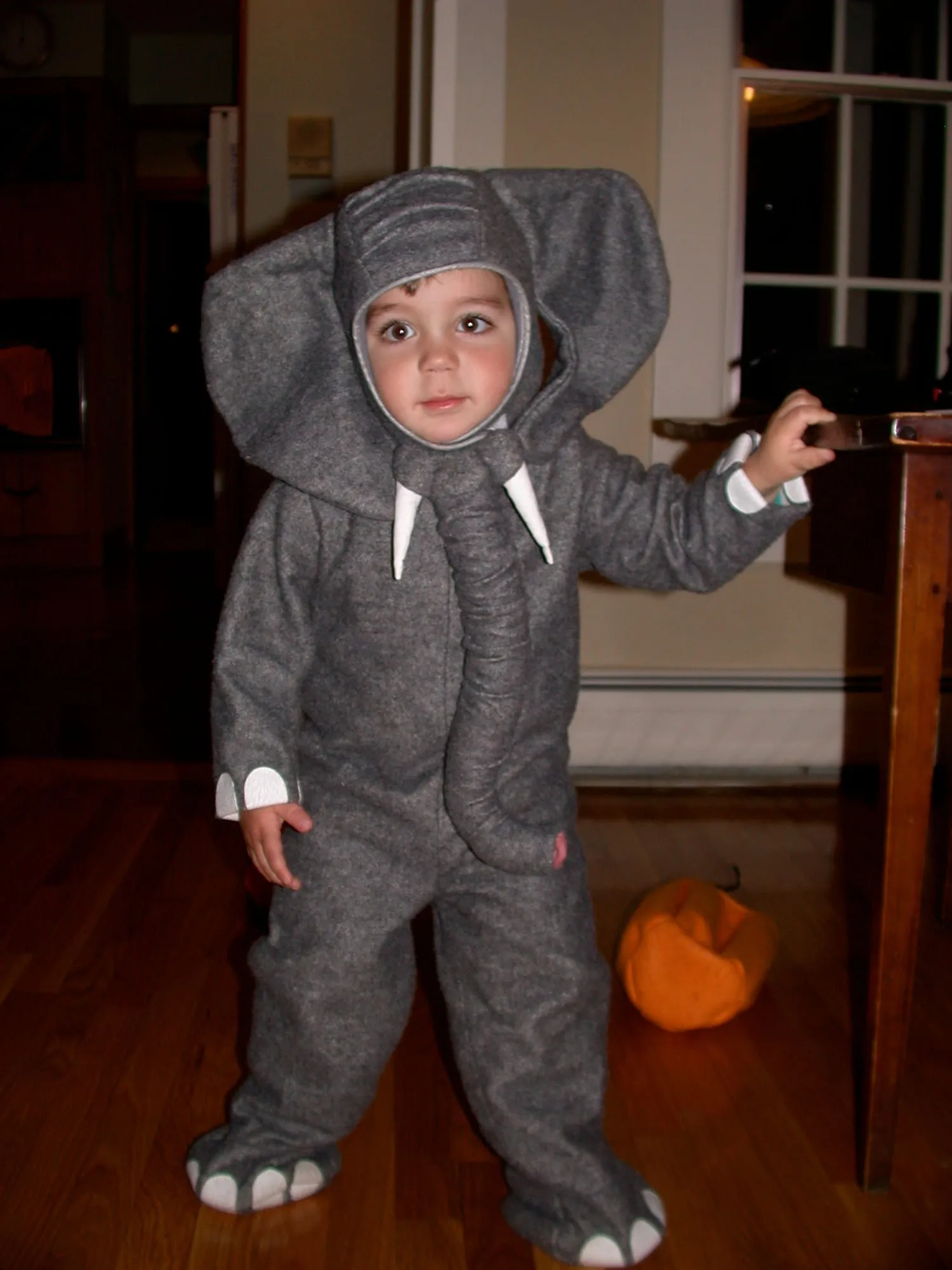 A toddler in a gray fleece elephant costume with large floppy ears, a sculpted hanging trunk, white felt tusks, and white toes on the feet.