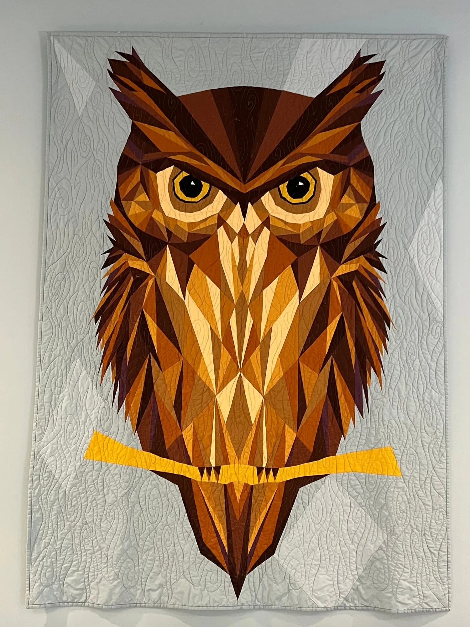 A quilt hung on a pale wall showing a great horned owl in browns, golds and cream, built from sharply angled pieced facets, gripping a yellow branch.