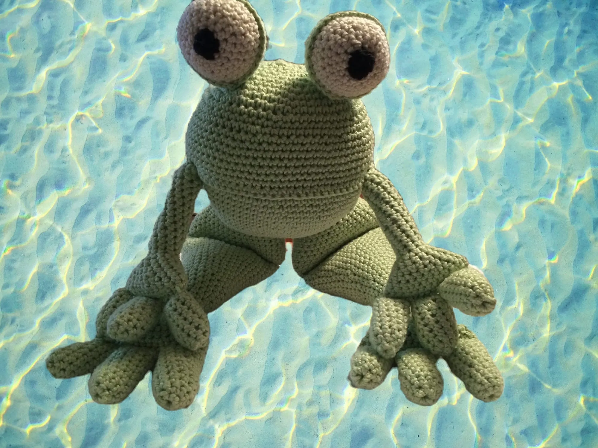 A crocheted frog with large round eyes and wide webbed feet, against a background of rippling blue water.