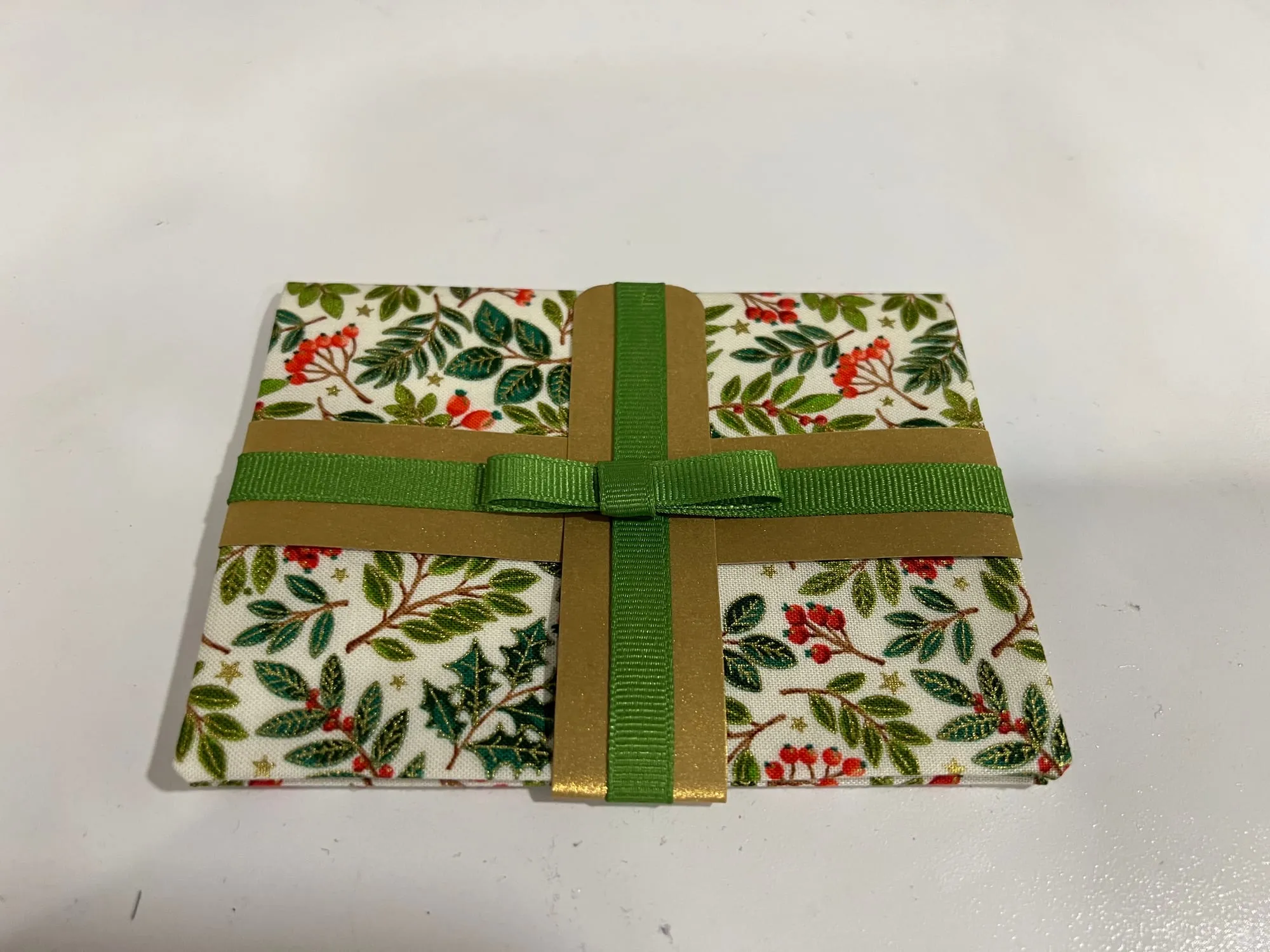 A greeting card covered in a holly and berry print, banded with a gold ribbon and a green ribbon tied in a flat bow.