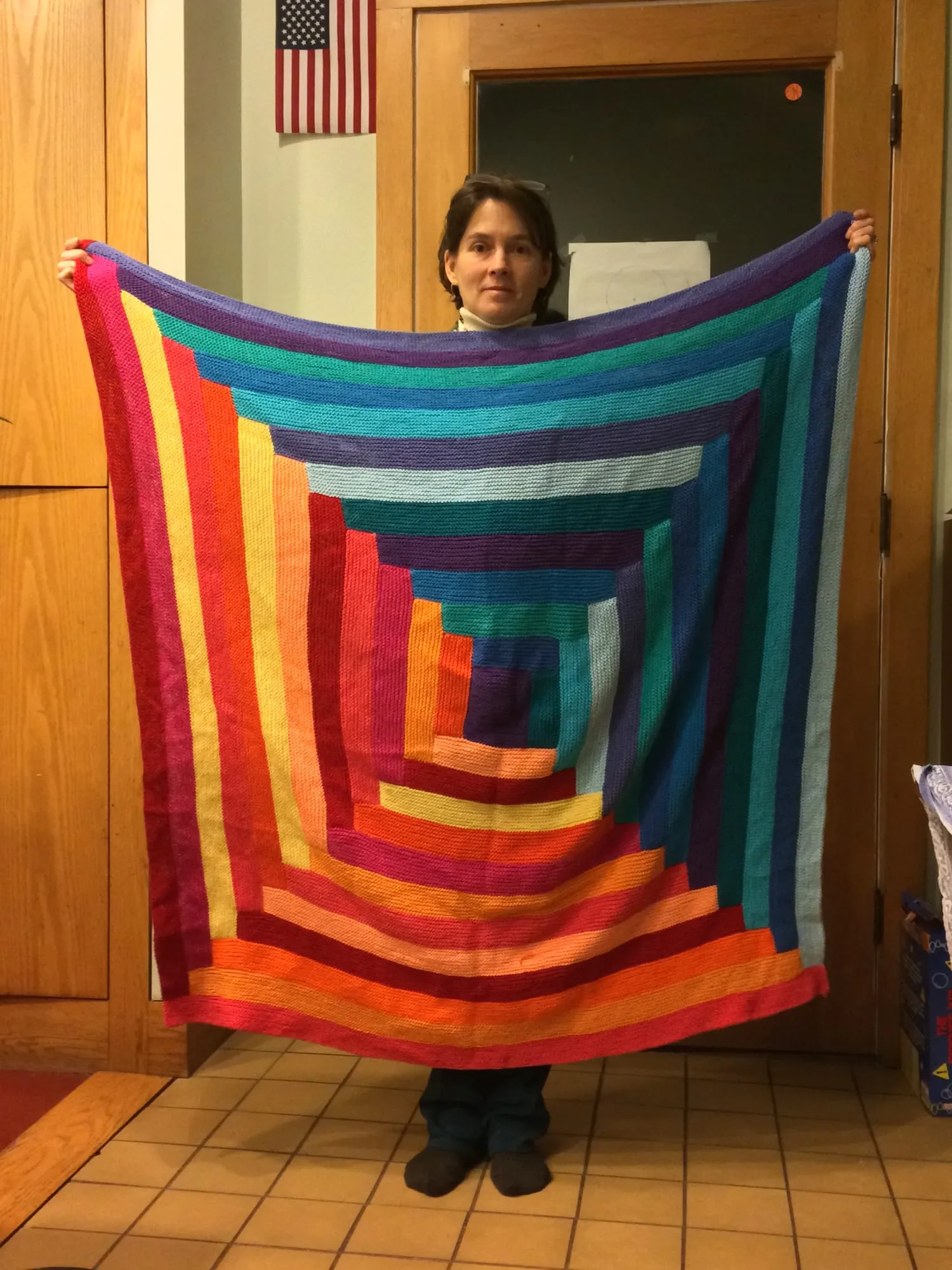 A knitted log cabin blanket, built outward from the center in rectangular rounds of red, orange, yellow, green, teal and purple, held up open at full width.