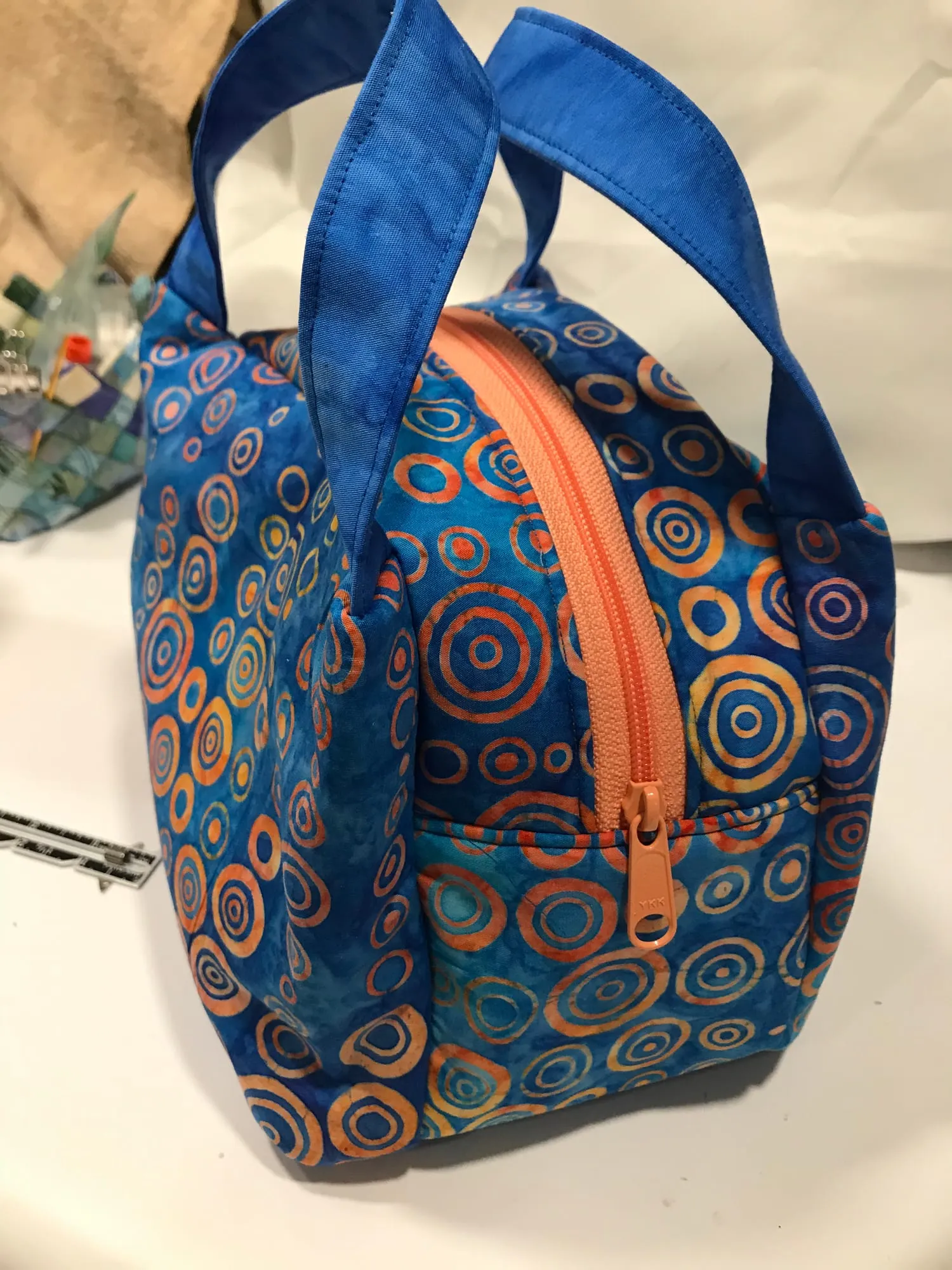 An insulated lunch bag in a blue fabric printed with orange and blue circles, with a coral zip and blue webbing handles.