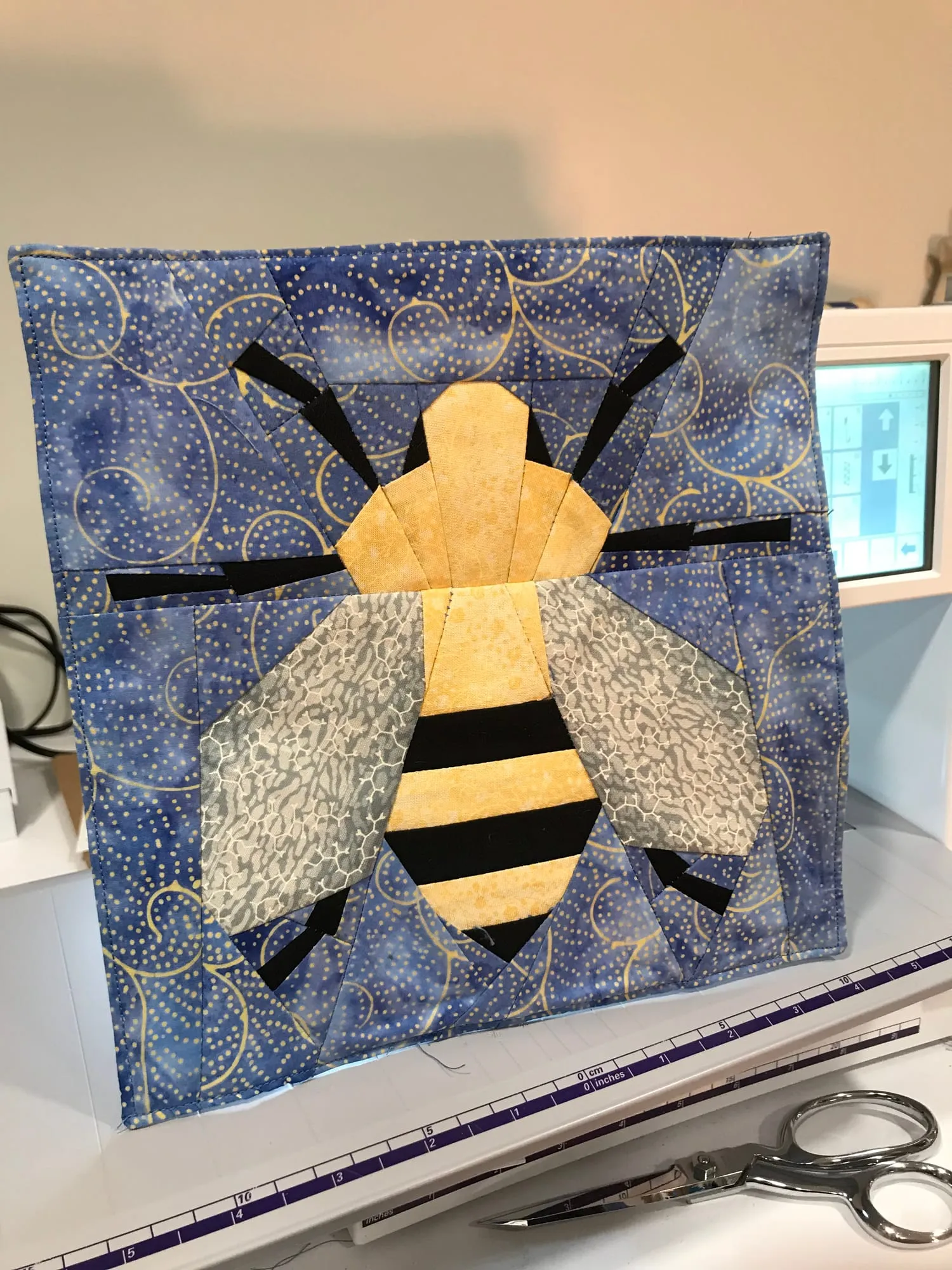 A small quilt propped upright on a sewing machine, showing an appliquéd honeybee with gold and gray wings on a blue printed ground.