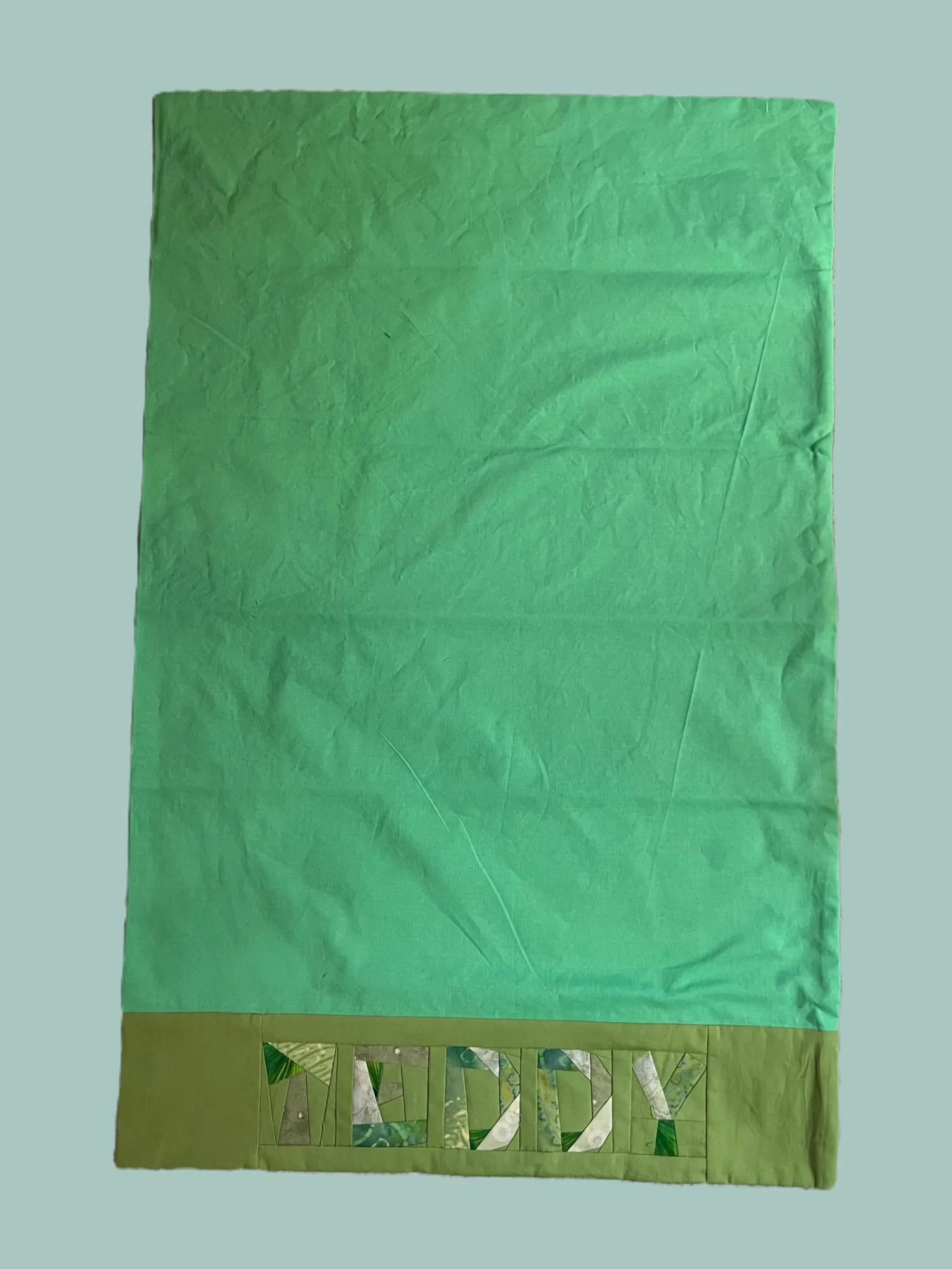 A green pillowcase with TEDDY across the band, each letter pieced from a different patterned green and white fabric.