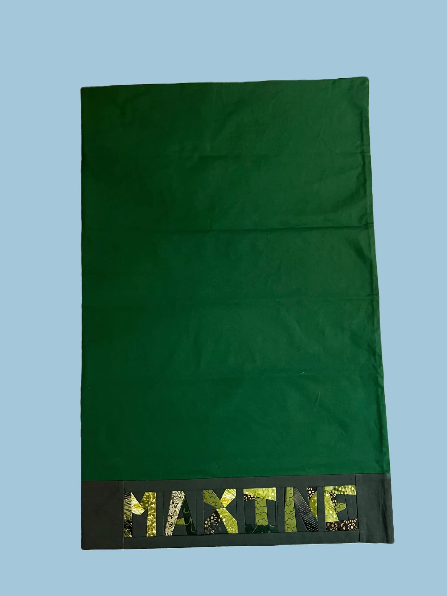 A dark green pillowcase with MAXINE across the band, each letter pieced from a different patterned green fabric.