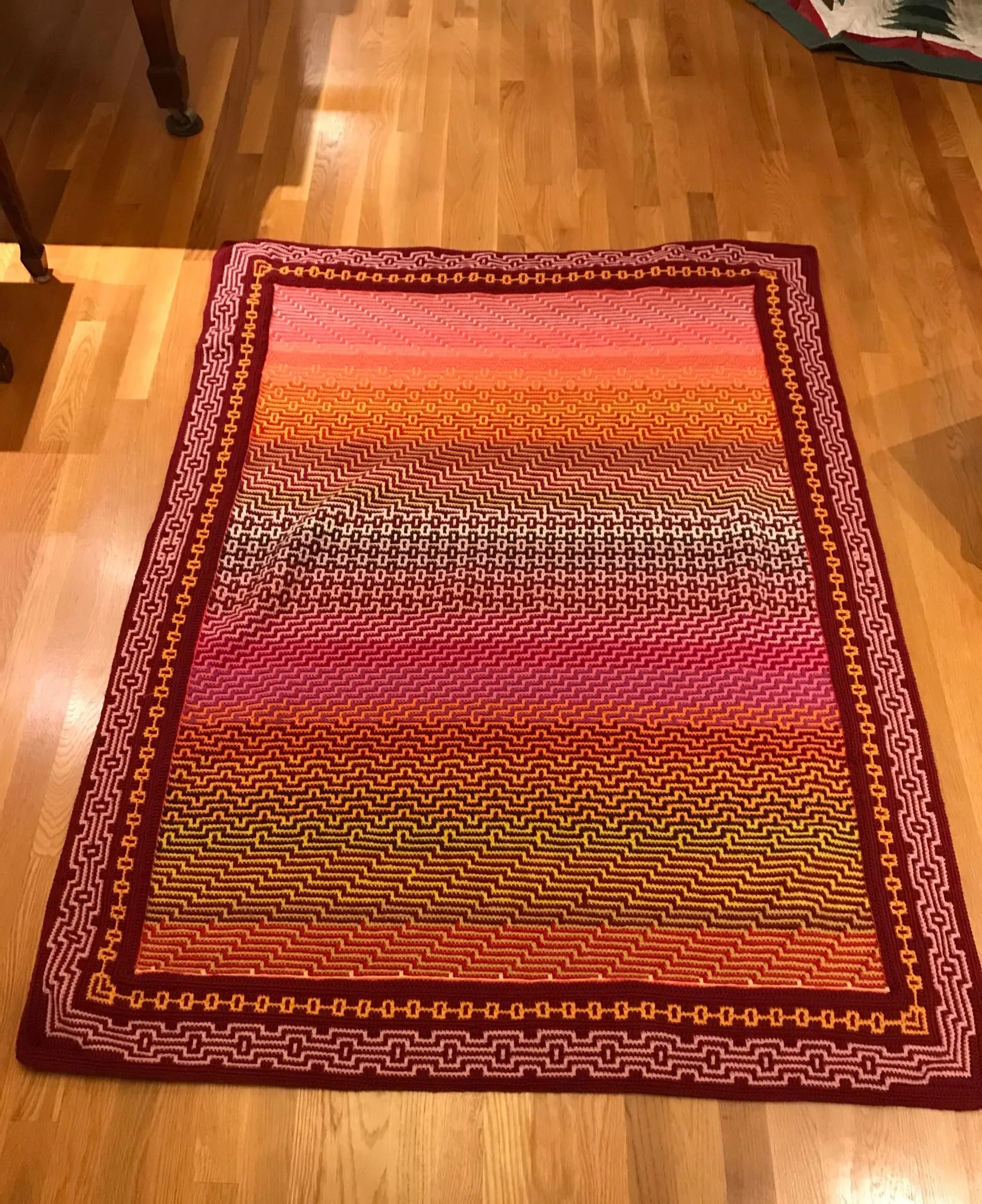 A mosaic crochet blanket on a wood floor, worked in rows that shade from deep red through orange to olive, inside a patterned dark red border.