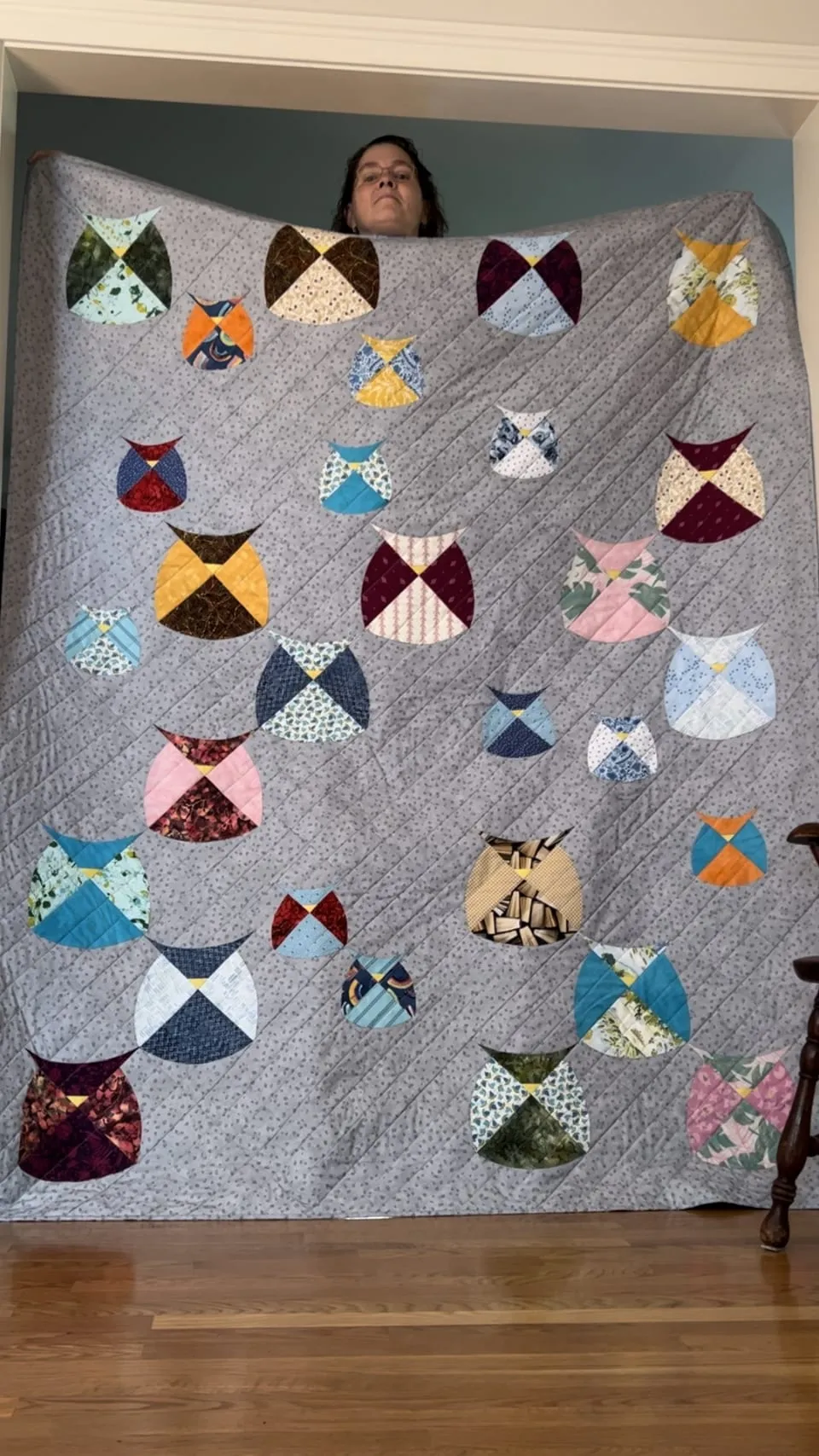 Sarah holding up a large gray quilt scattered with pieced owls in many different fabrics, each owl a circle split into colored quarters with a small yellow beak, her face visible above the top edge.