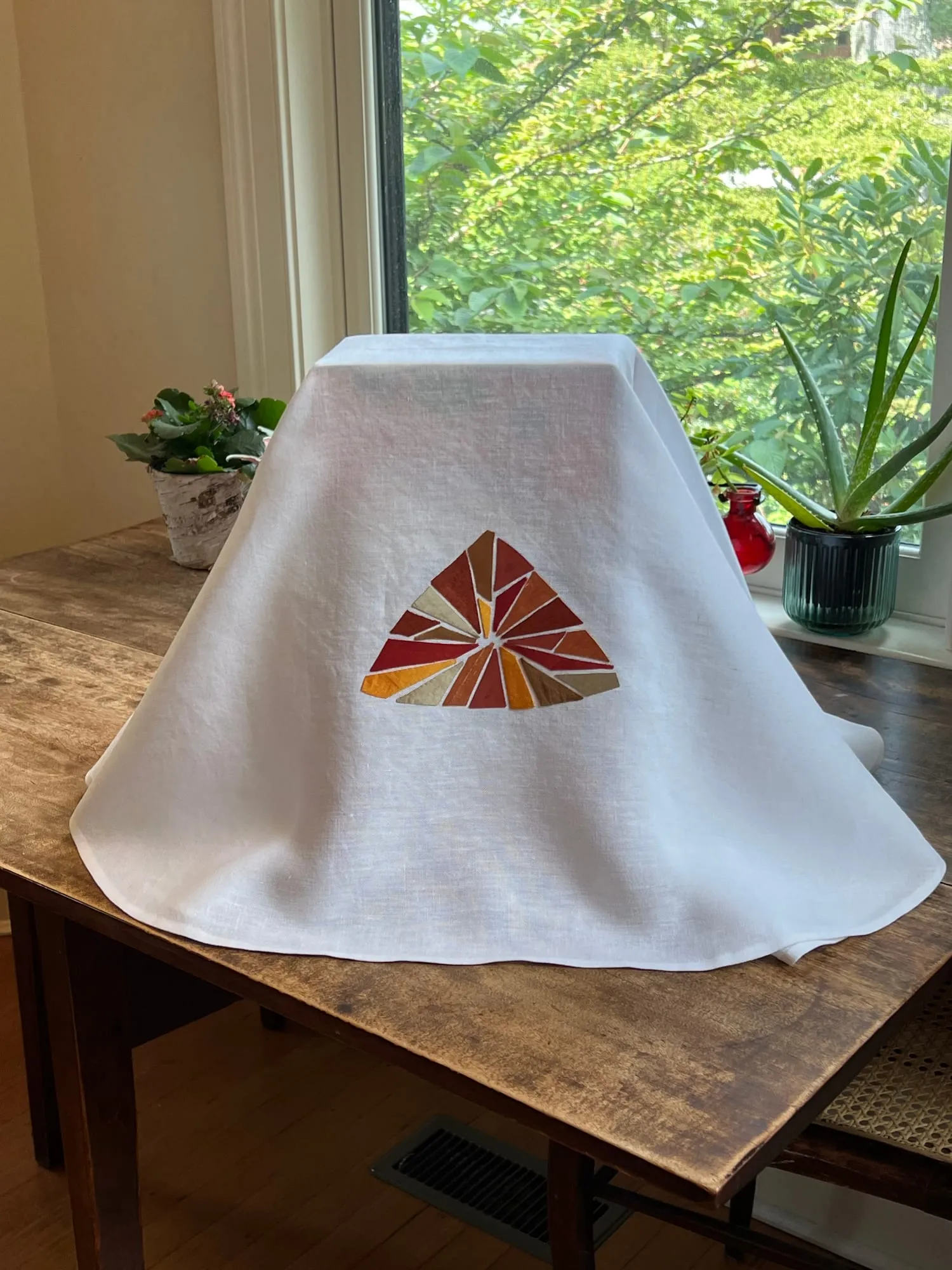 A white pall draped over a table, with a small triangular appliqué at the center in gold, red and brown.