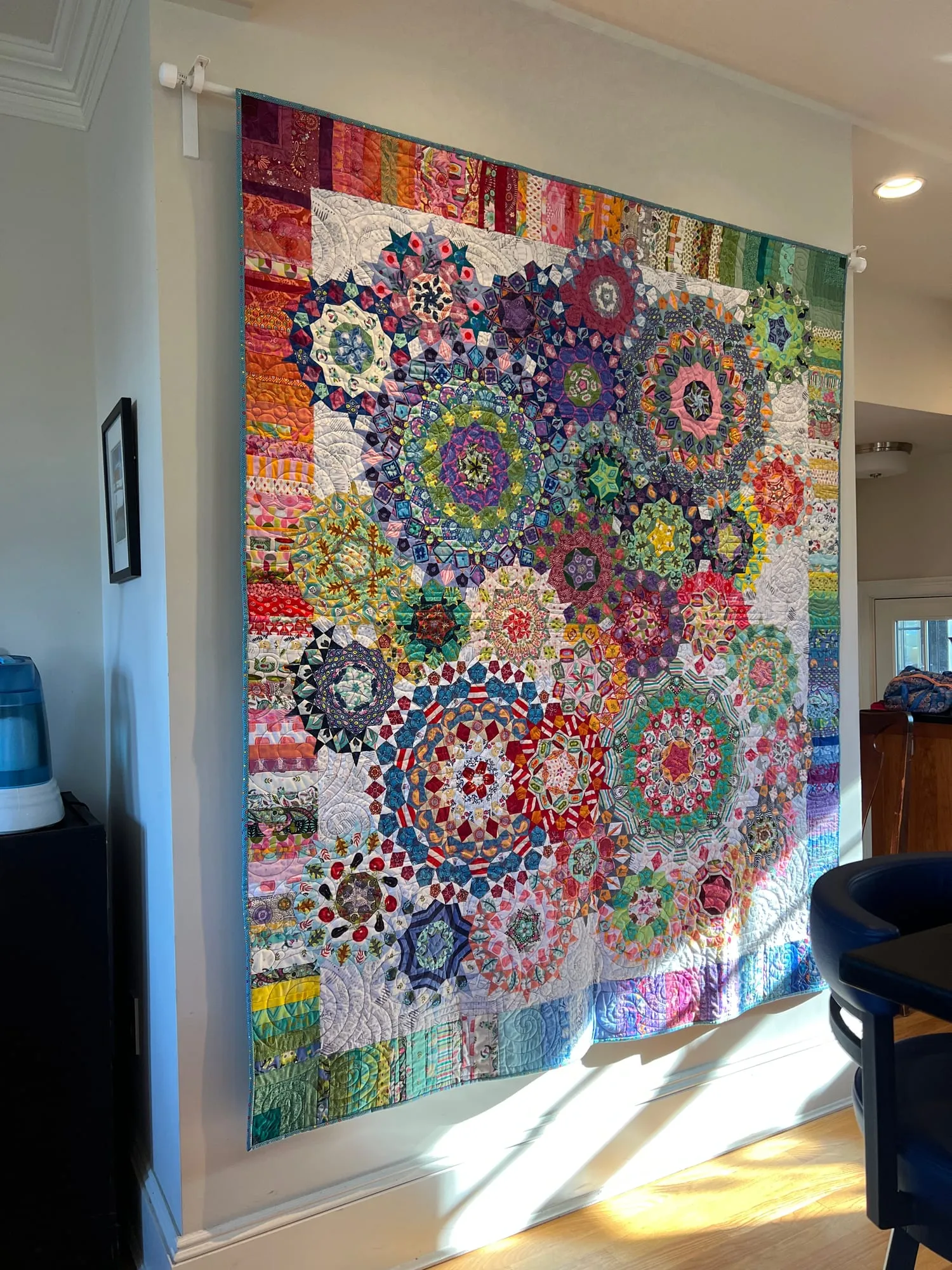 A large quilt hung against a bright window wall, pieced from hundreds of small printed hexagons and diamonds into interlocking rosettes in every color, on a white ground.
