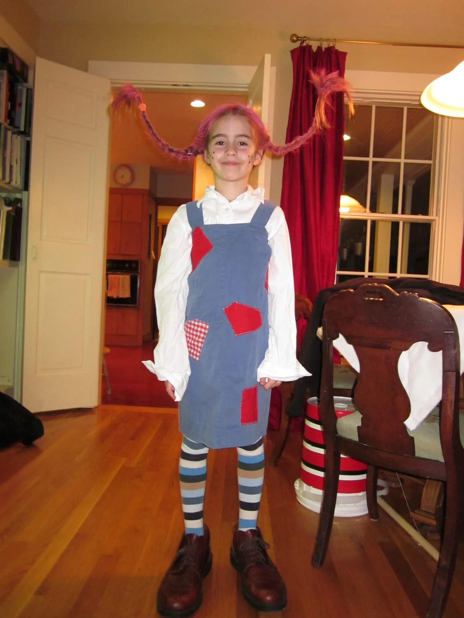 A child dressed as Pippi Longstocking in a blue patched pinafore over a white men's button-down shirt, with striped knee socks, brown boots and braided red-pink plaits.