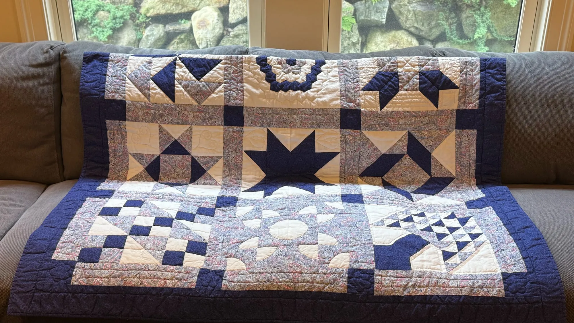 A patchwork watercolor quilt in navy, paisley purples and white spread over a sofa, pieced in blocks: eight-pointed stars, pinwheels and nine-patch squares, inside a deep navy border. All hand quilted.