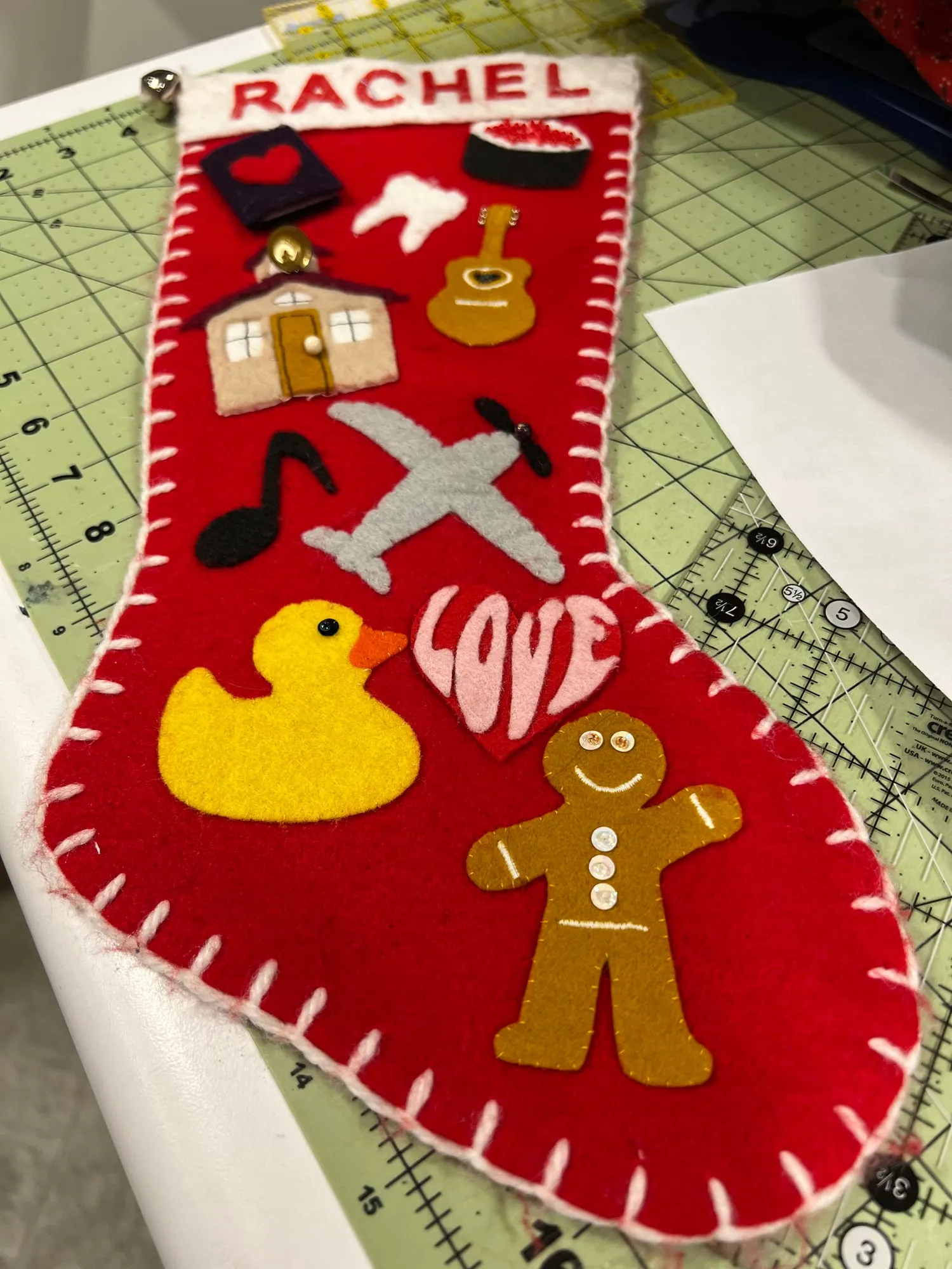 A red felt Christmas stocking edged in white blanket stitch, with RACHEL appliquéd across a white cuff, and shapes down the front including a book, a tooth, a house, a guitar, music notes, an airplane, a yellow duck, a heart reading LOVE and a gingerbread man.