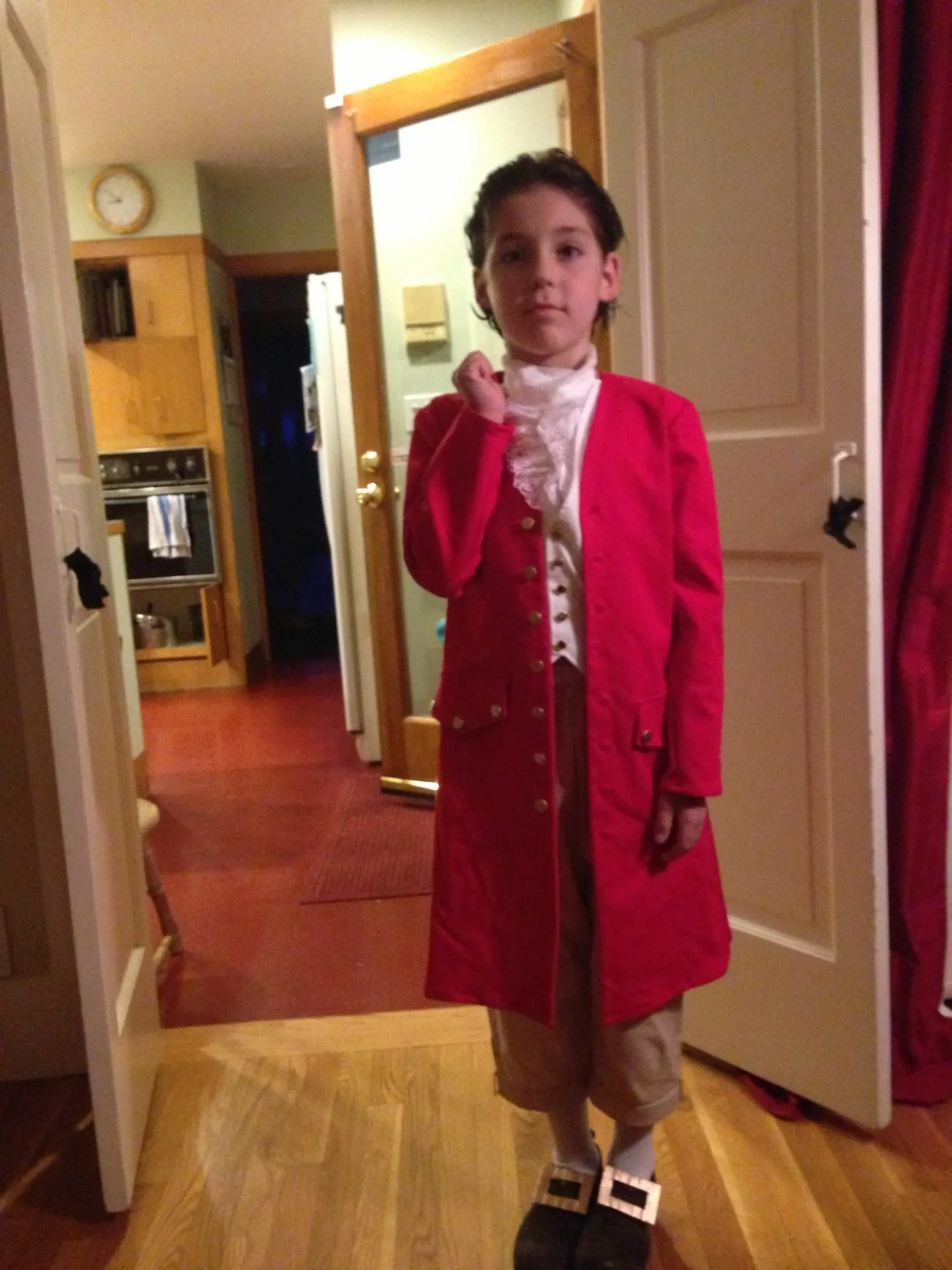 A child in a long red colonial coat with a row of round buttons, worn over a white shirt with a ruffled jabot, with tan breeches, white stockings and buckled shoes.