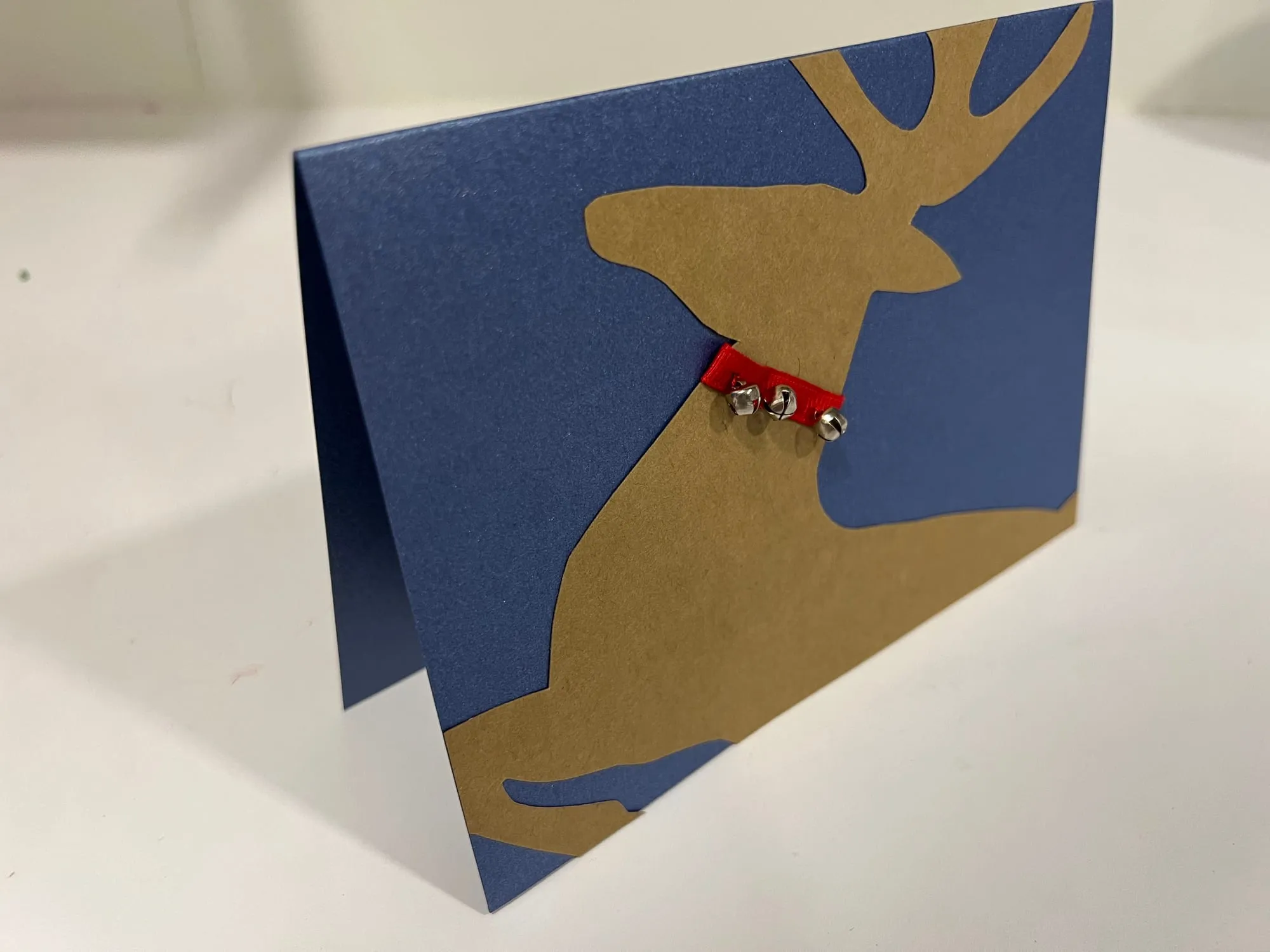 A navy blue card with a gold-brown reindeer silhouette on the front, wearing a red collar strung with small bells.