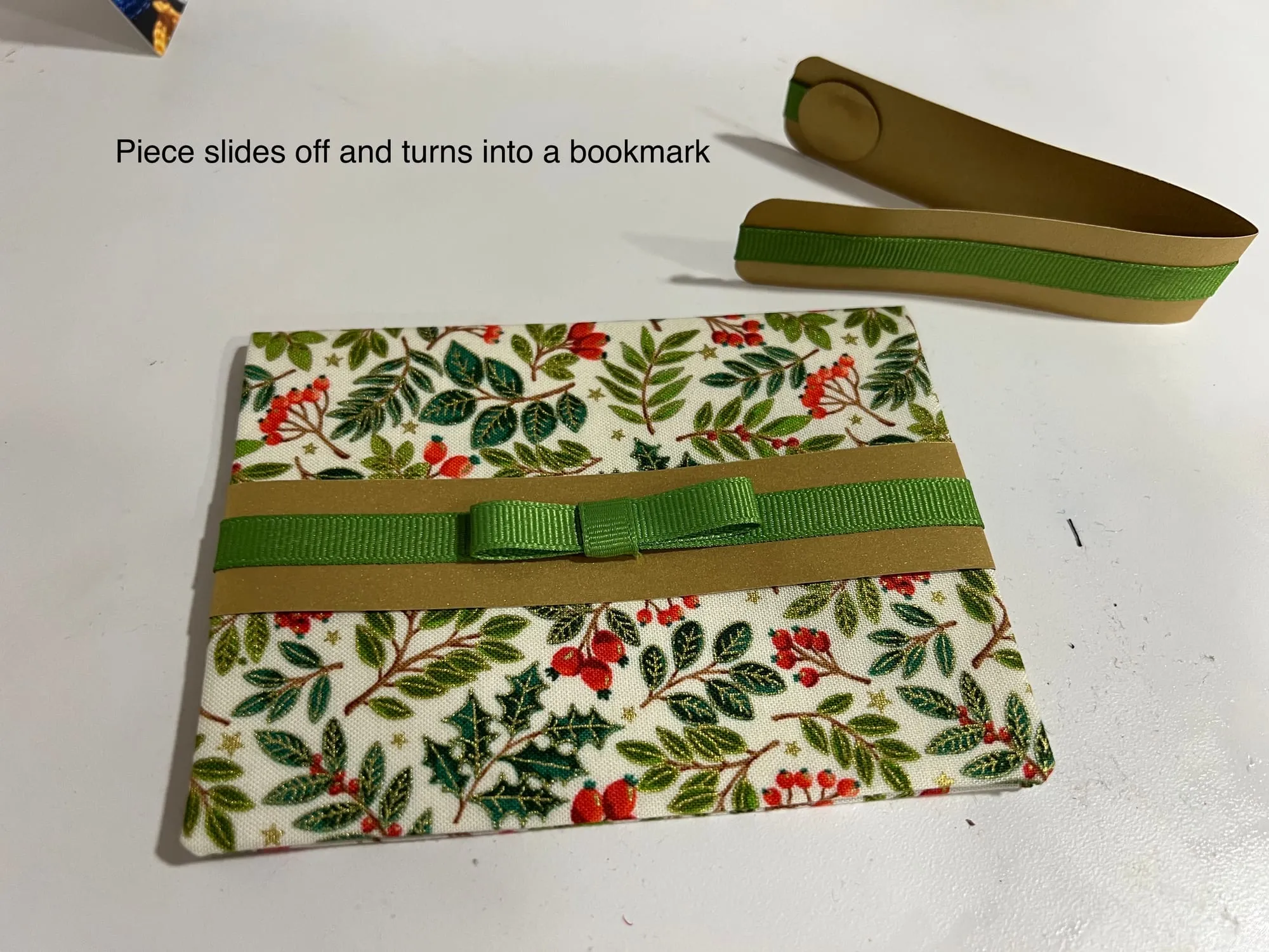 The same holly-print card with its ribbon band slid off and lying beside it, showing the piece that becomes a bookmark.