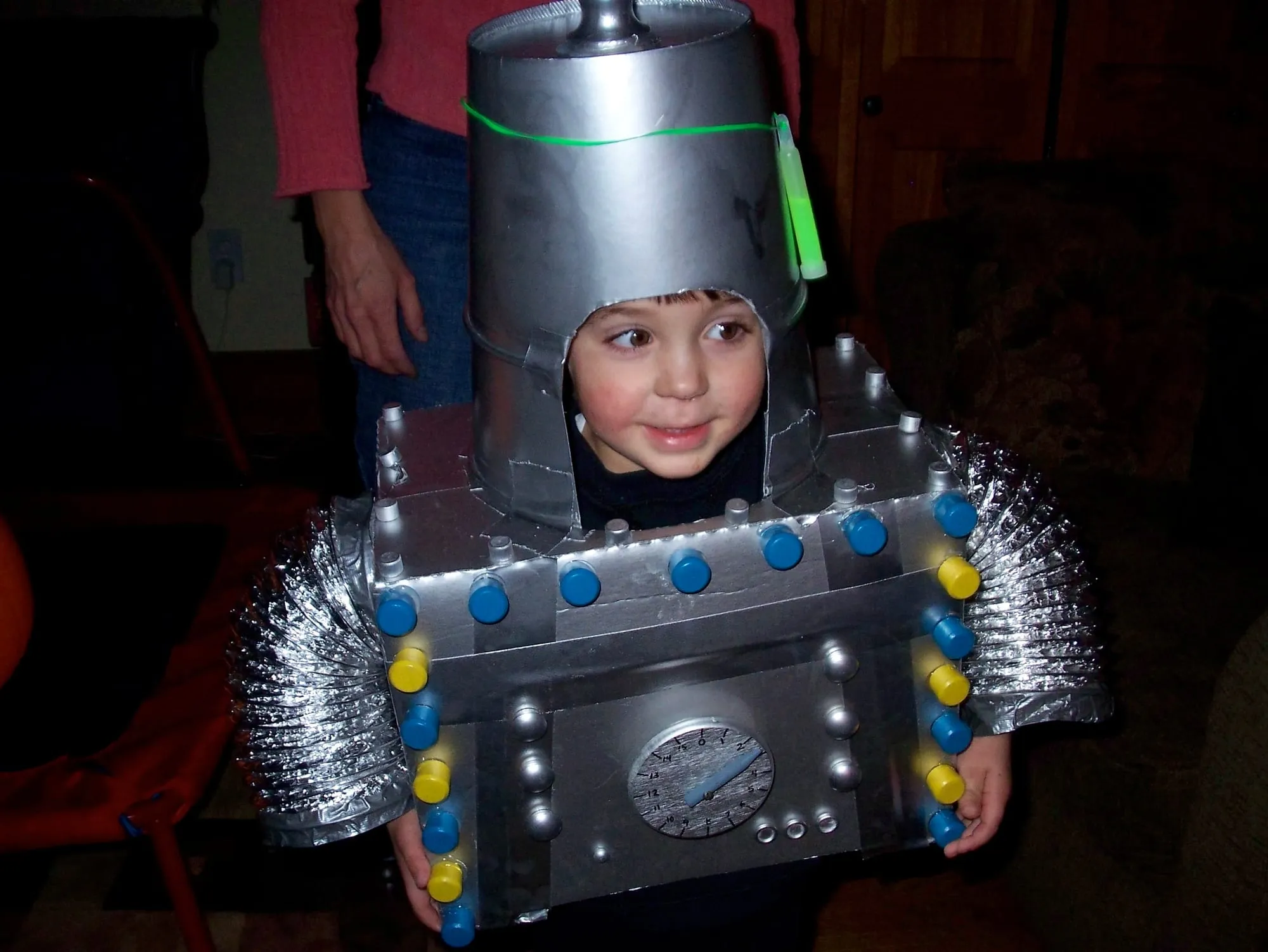 A child in a silver robot costume built from a boxy body panel studded with blue and yellow bottle caps, a dial gauge on the front, ducting for arms, and a bucket helmet with a glow stick.