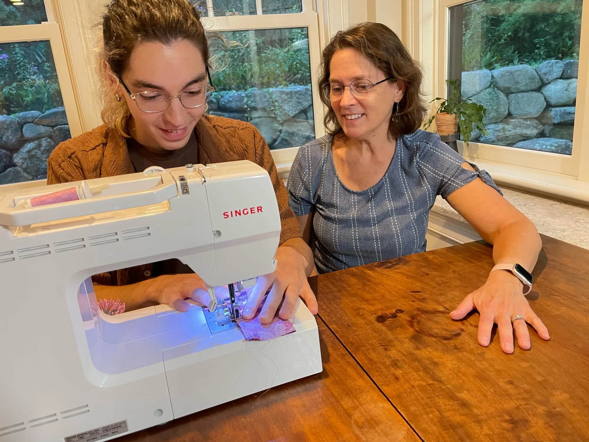 A young man sewing a sampler on a Singer machine, both hands guiding the fabric under the needle, with Sarah sitting beside him watching the stitching.
