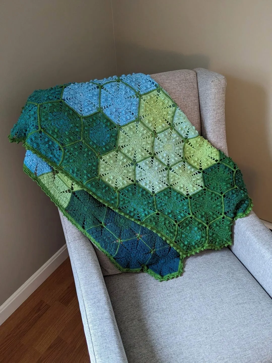 A crocheted hexagon blanket shading from deep green through pale green to blue, draped over the arm of a pale armchair.