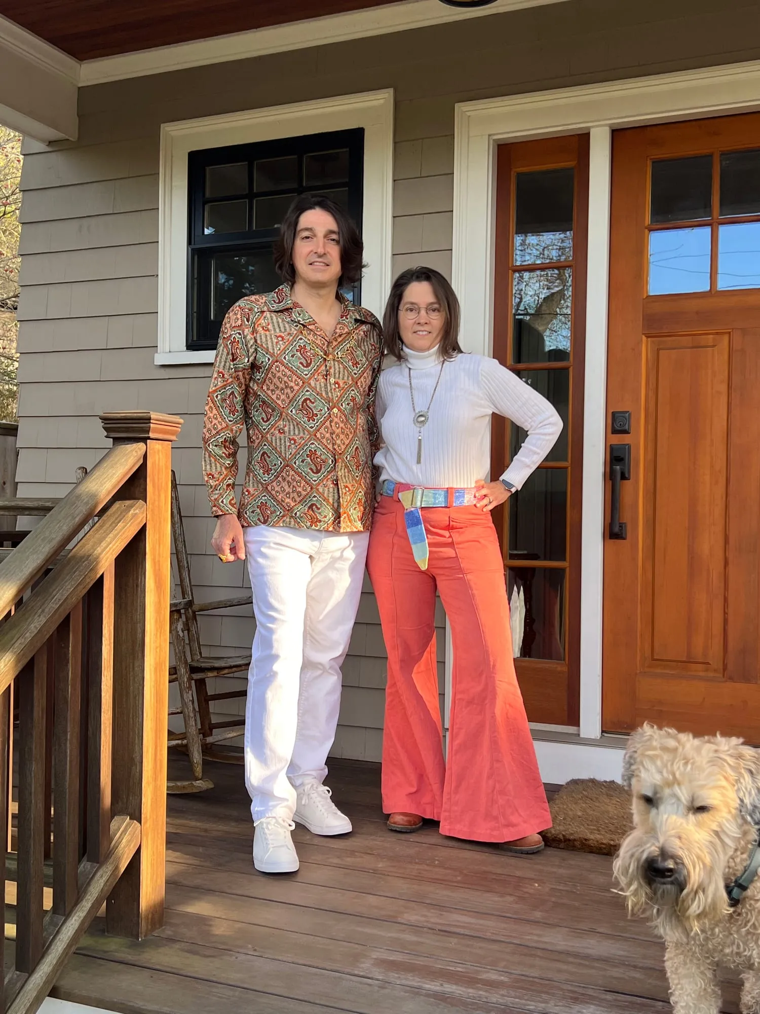 Two adults on a porch dressed for a seventies party, orange flared trousers with a patchwork belt worn with a white turtleneck, beside a patterned shirt and white trousers.