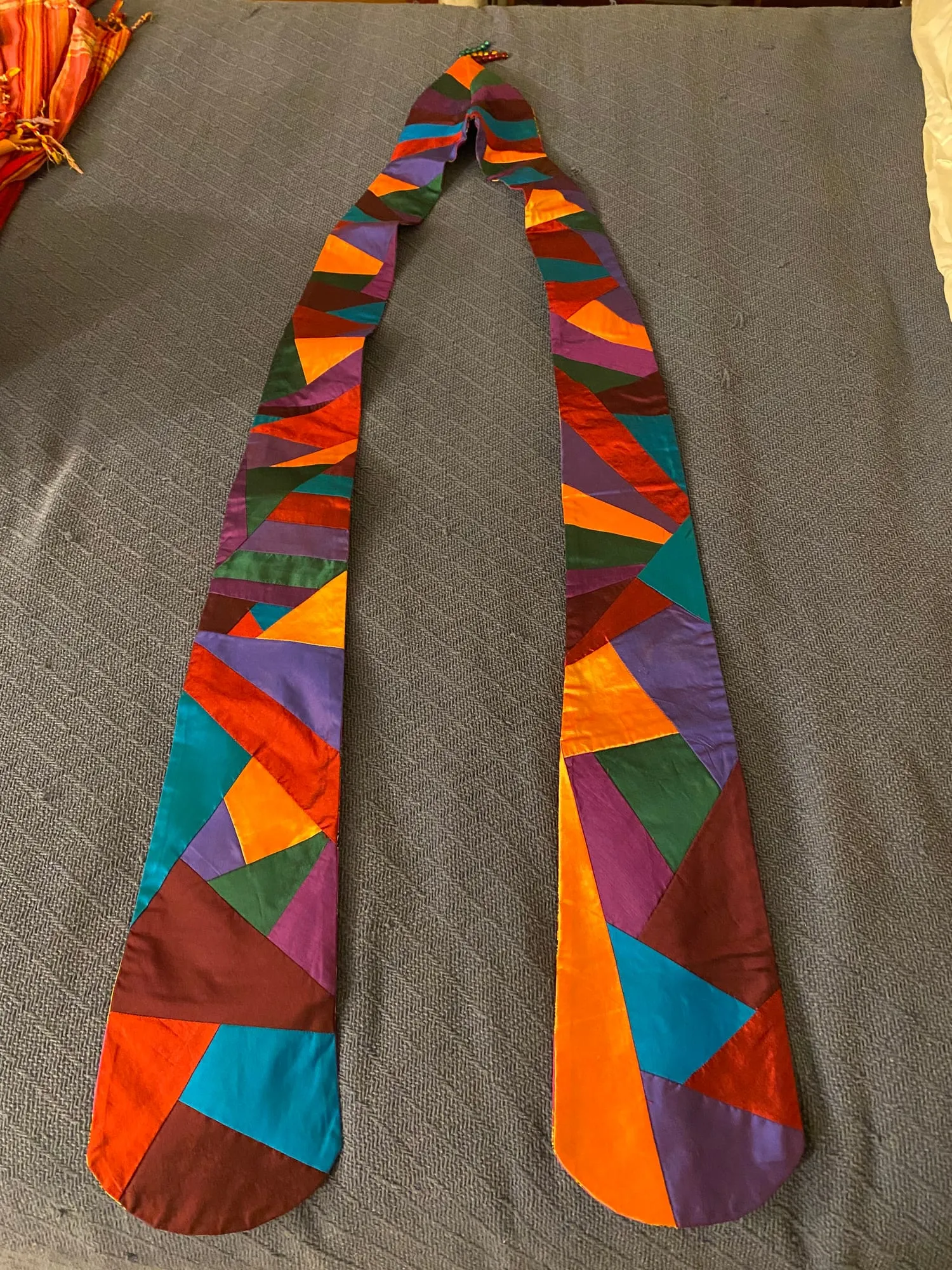 A silk stole laid flat on gray cloth, pieced from irregular blocks of orange, red, teal, purple and brown fitted together like stained glass.