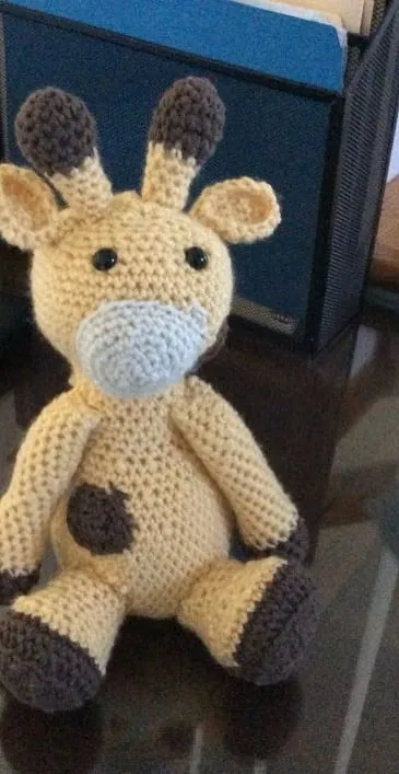 A crocheted giraffe in pale cream with brown horns, hooves and spots, sitting on a glass tabletop.