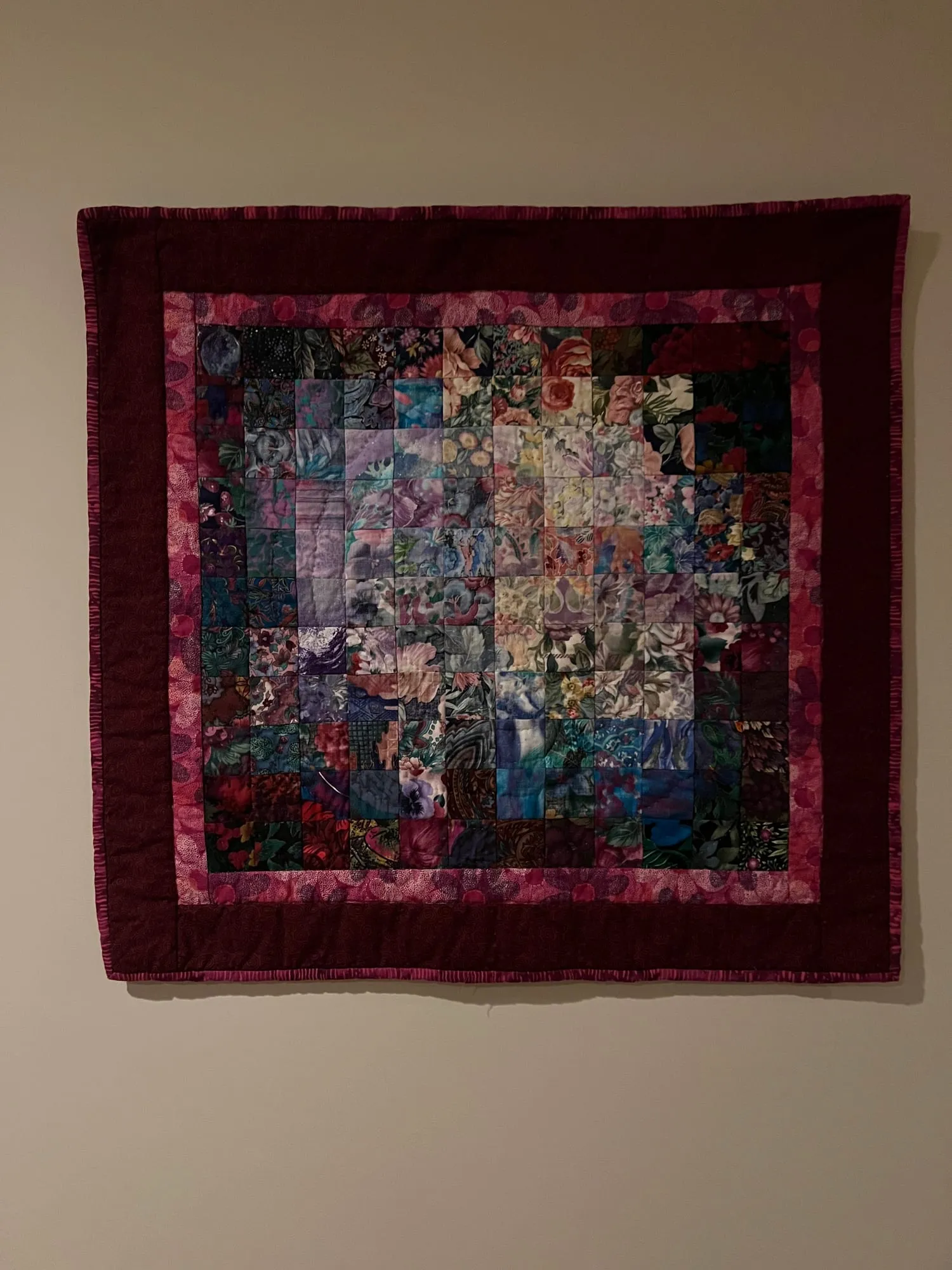 A small quilt hung on a wall, its center pieced from hundreds of tiny dark floral squares, framed by borders in magenta and deep red.