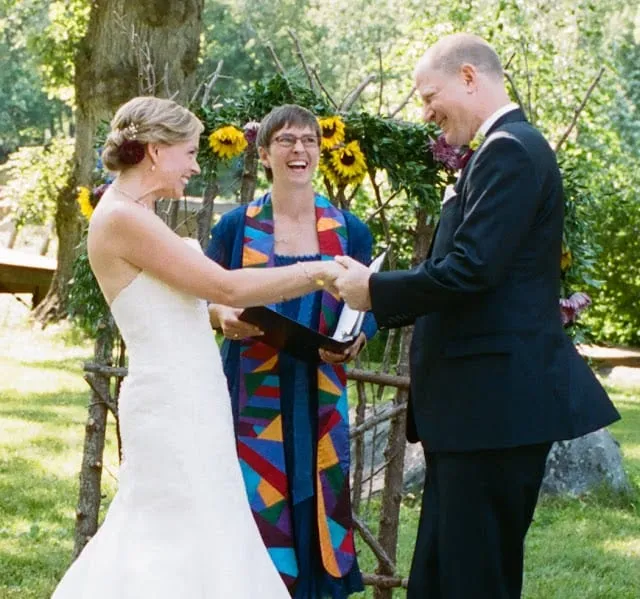 A bride and groom holding hands at an outdoor ceremony, the officiant standing between them in a brightly pieced stole.